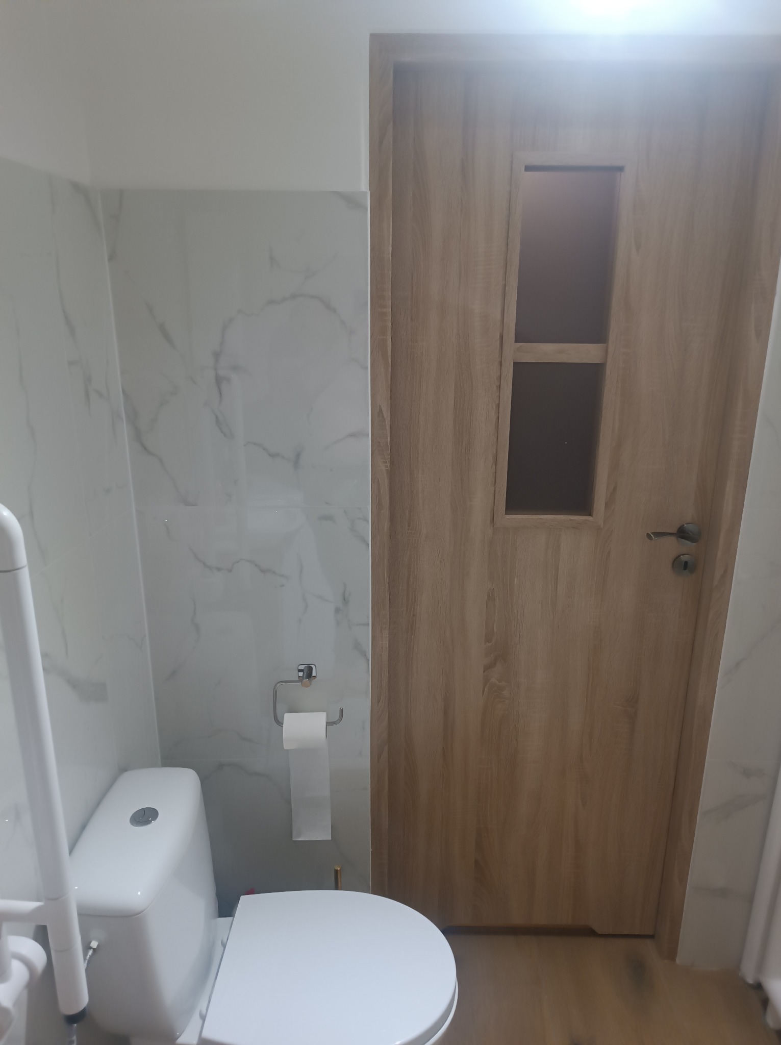 Biała toaleta z uchwytem dla niepełnosprawnych, marmurowe płytki ścienne i drewniane drzwi z szybą w łazience po remoncie. Widok z niskiego kąta.
