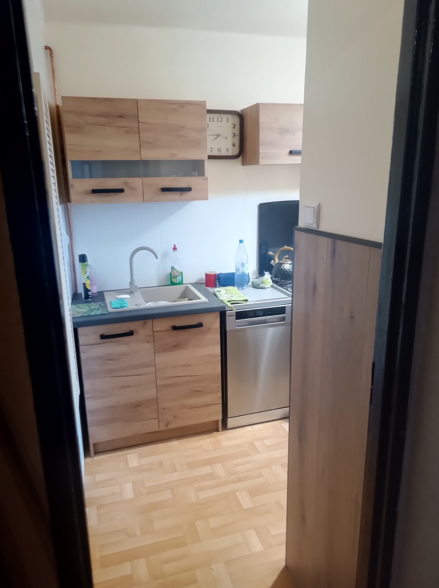 Ujęcie z perspektywy wejścia do kuchni. Widoczne drewniane szafki, zlewozmywak, srebrna zmywarka i parkiet. Na ścianie zegar i elementy wyposażenia.
