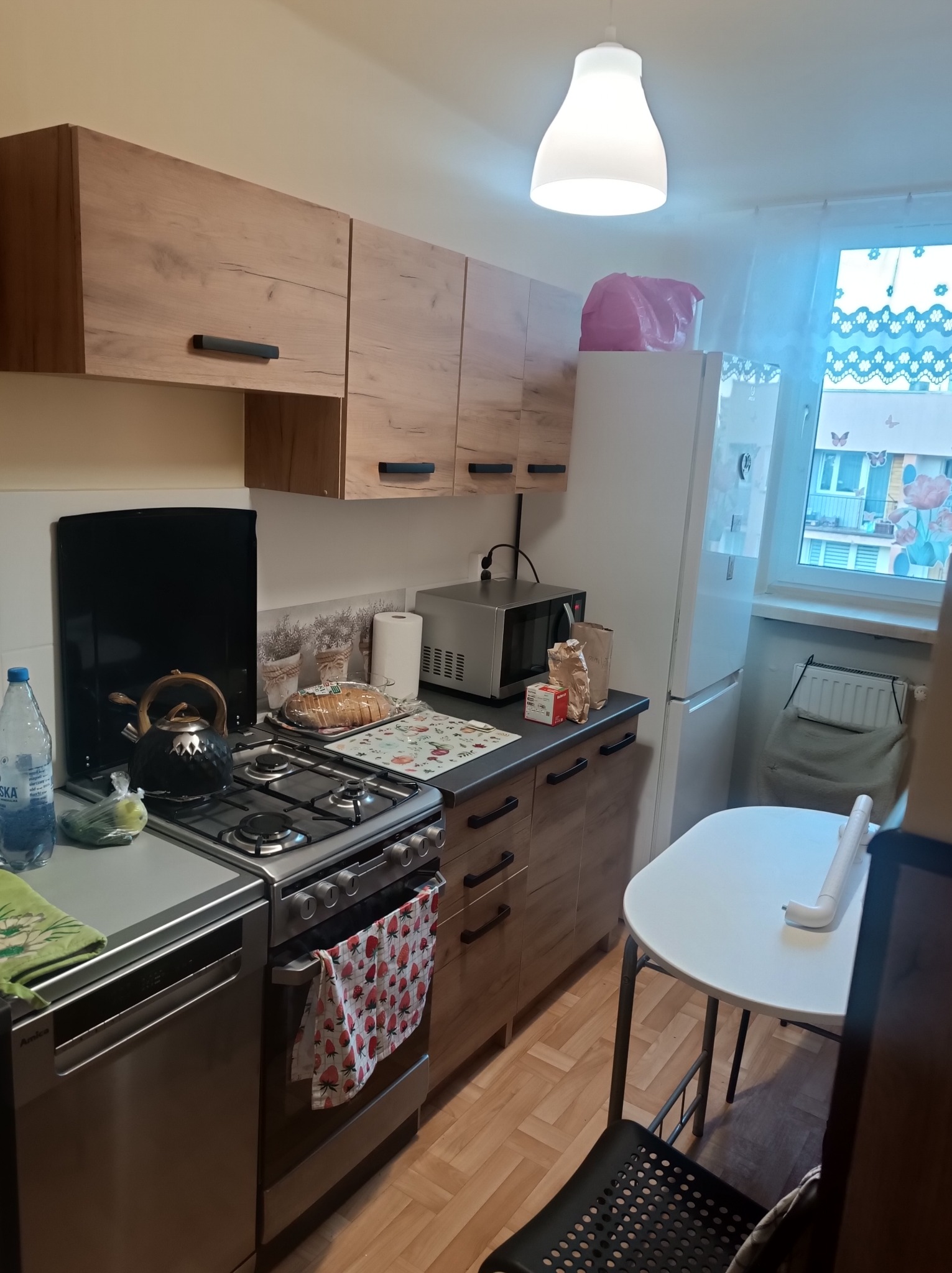 Funkcjonalna, choć niewielka kuchnia z drewnianymi szafkami, srebrną kuchenką i lodówką. Na blacie czajnik, mikrofala i drobne akcesoria. Widok na okno z firanką.