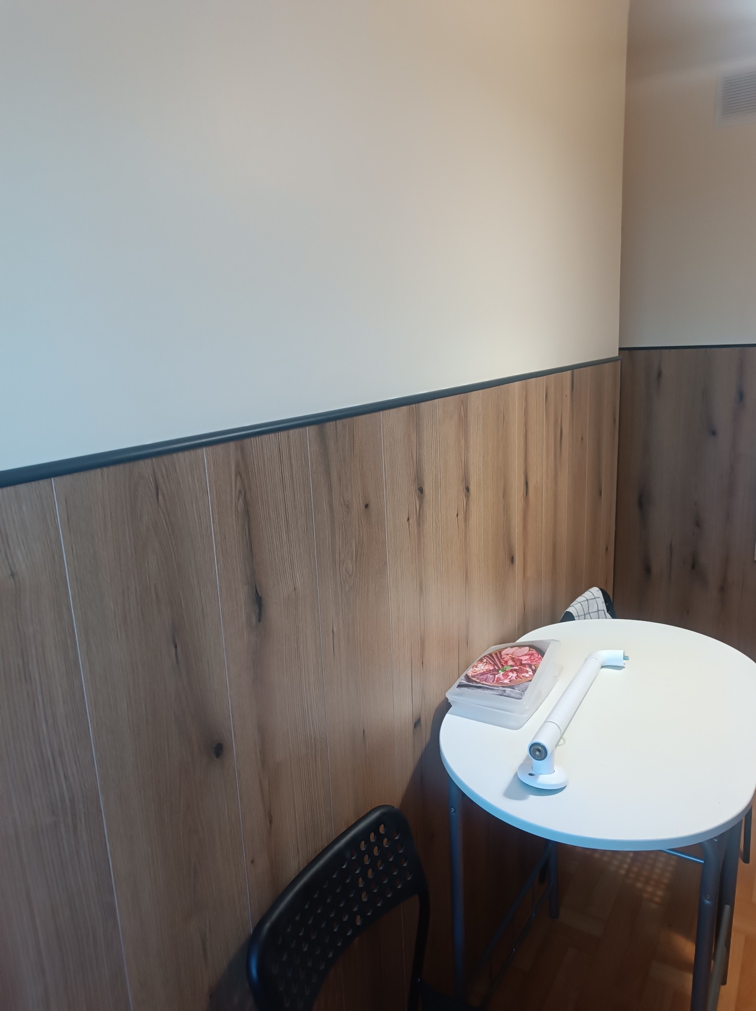 Panel ścienny w kolorze drewna z czarną listwą, biała ściana powyżej. Na stole pizza i lampa. Czarny fotel w rogu. Wykończenie wnętrza.