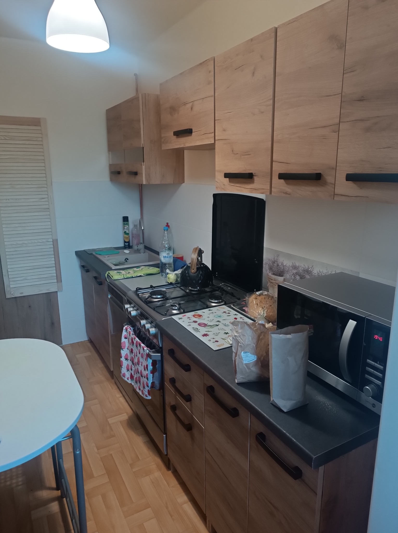 Jasna kuchnia z drewnianymi szafkami i czarnymi uchwytami, kuchenka gazowa, mikrofalówka i biały stół. Widoczne akcesoria kuchenne i płytki imitujące drewno.