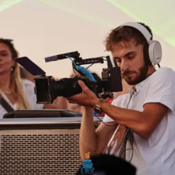 Młody mężczyzna w słuchawkach, ubrany w białą koszulkę polo, obsługuje profesjonalną kamerę filmową z osprzętem na tle rozmytych postaci i scenicznego oświetlenia.