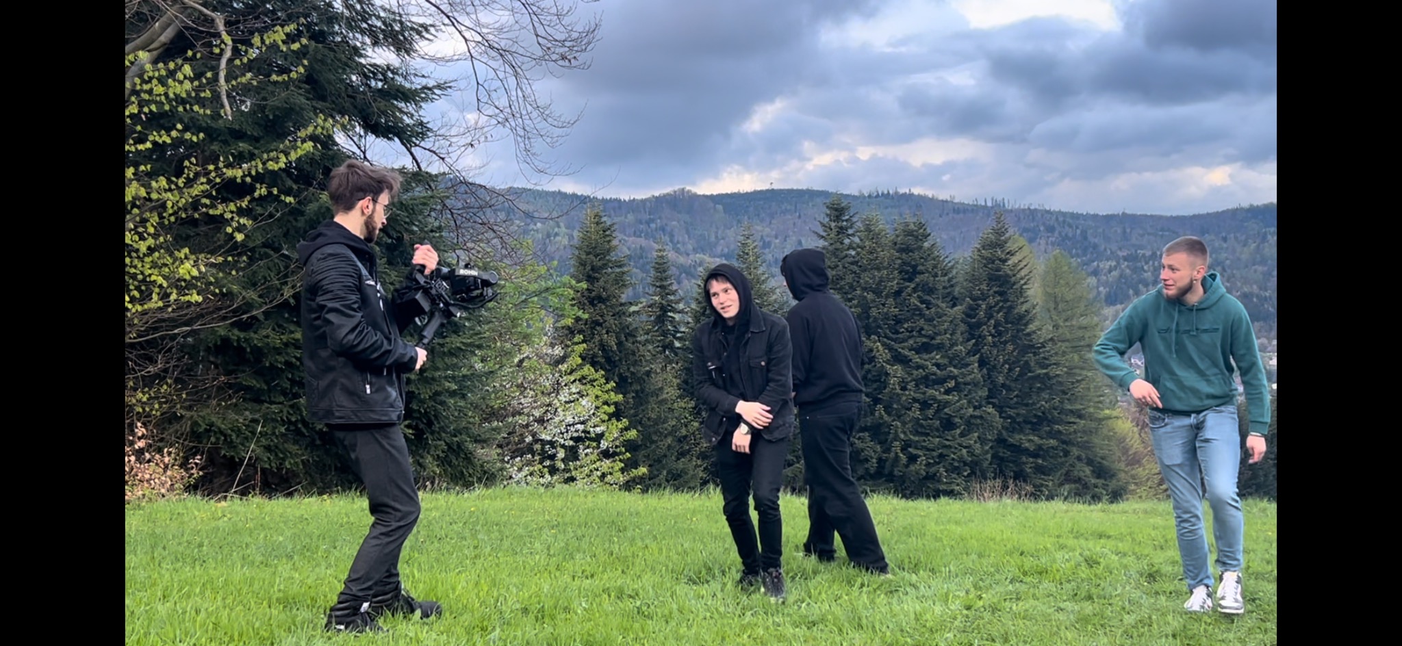 Ekipa filmowa kręci scenę w plenerze: kamerzysta z profesjonalnym sprzętem, dwóch aktorów w czarnych strojach i model w zielonej bluzie na tle górskiego krajobrazu.