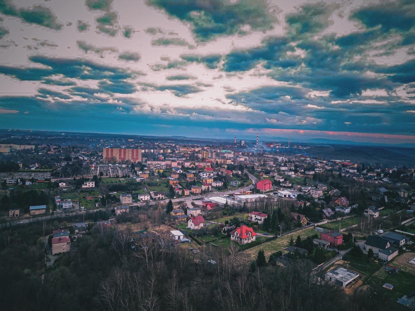 Panoramiczny widok z drona na miasto o zmierzchu, z widocznymi budynkami mieszkalnymi, terenami zielonymi, kominami fabrycznymi w oddali i dramatycznym, zachmurzonym niebem.