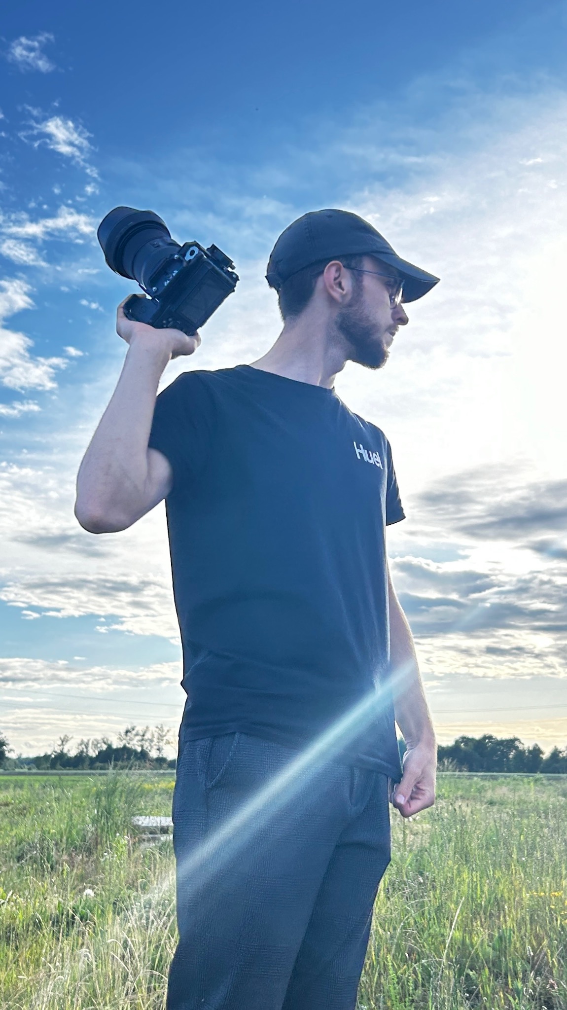 Mężczyzna w czarnej koszulce i czapce z daszkiem, trzymający w dłoni aparat fotograficzny na tle nieba z chmurami i łąki.