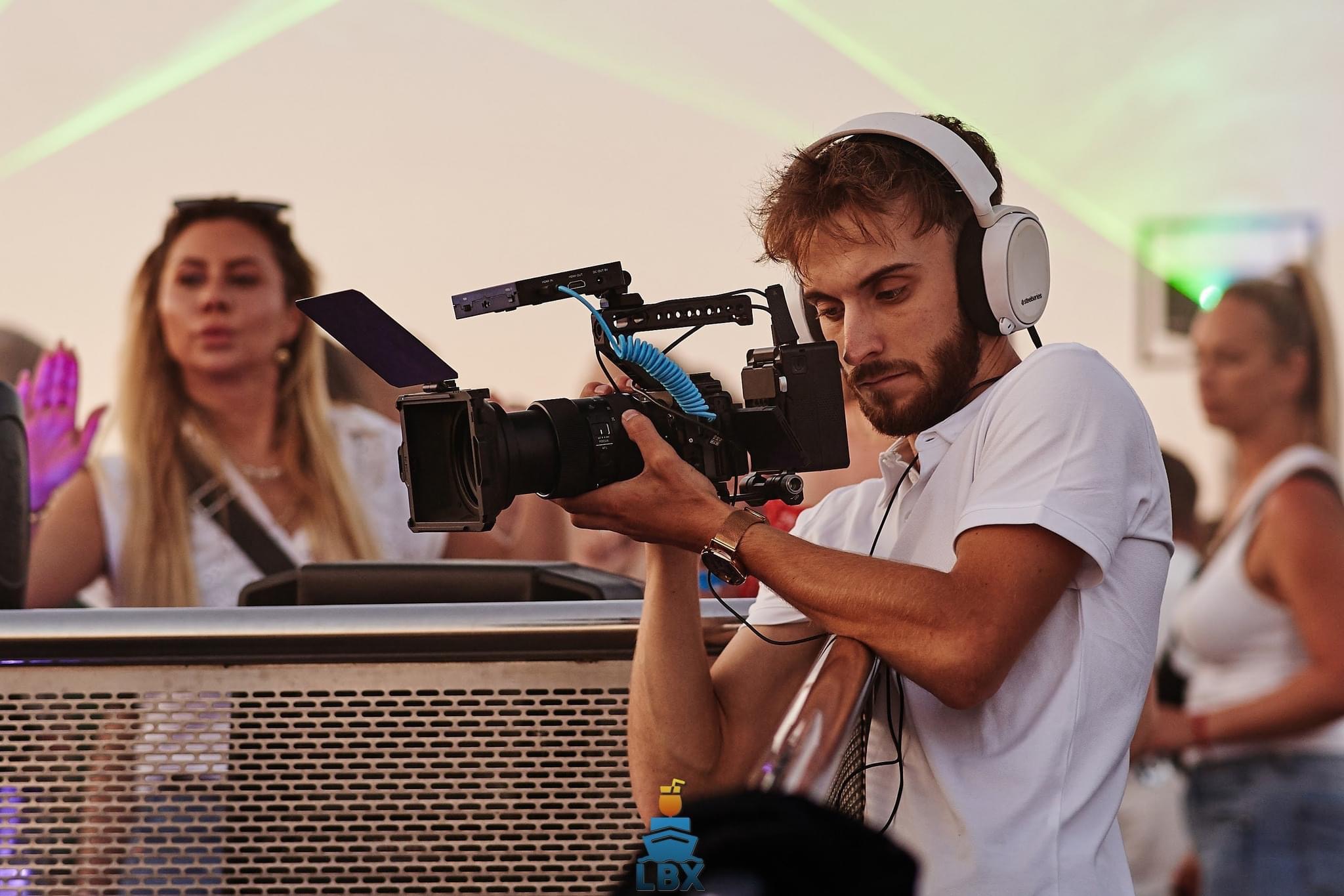 Młody mężczyzna w słuchawkach, ubrany w białą koszulkę polo, obsługuje profesjonalną kamerę filmową z osprzętem na tle rozmytych postaci i scenicznego oświetlenia.