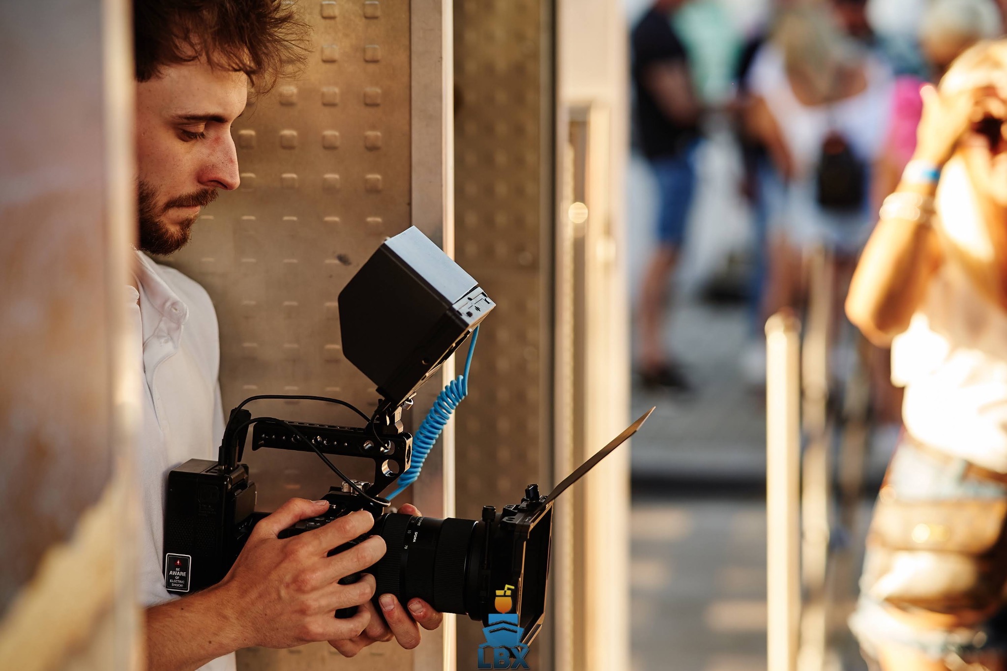 Młody mężczyzna z brodą obsługuje profesjonalną kamerę filmową z osprzętem, w tle rozmyte postacie na zewnątrz.