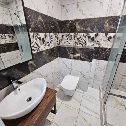 Natalia Meliakh Meliastrong - Nowoczesna łazienka z marmurowymi płytkami w odcieniach bieli i czerni, wzbogacona o dekoracyjny pas z motywem liści. Widoczna umywalka nablatowa na drewnianej szafce, toaleta oraz kabina...