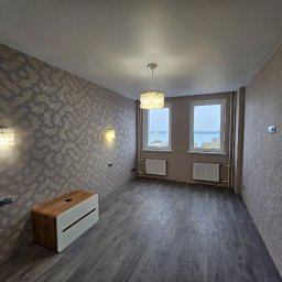 Natalia Meliakh Meliastrong - Wnętrze pokoju po remoncie: ciemne panele podłogowe, wzorzysta tapeta w odcieniach szarości i beżu, dekoracyjne lampy ścienne i sufitowa, widok na jezioro z okien.