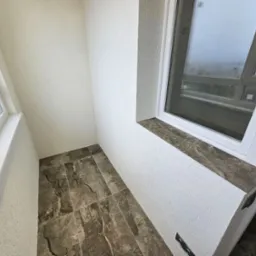 Wnętrze balkonu z szarobrązową marmurkową podłogą, białe ściany z fakturą baranka i białe okno z marmurowym parapetem.