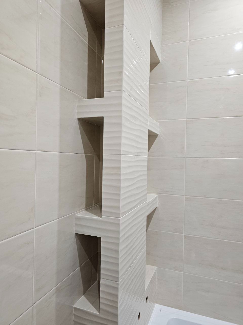 Wykończona łazienka z beżowymi płytkami i wbudowanymi, podświetlonymi półkami w pionowej kolumnie z falistym wzorem, widoczny fragment wanny.