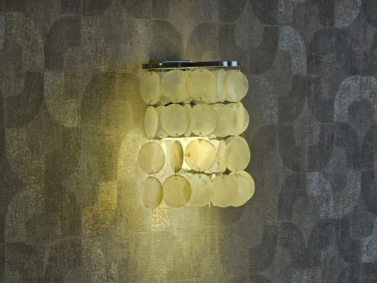 Kinkiet z okrągłymi, perłowymi elementami na tle ściany z geometrycznym wzorem w odcieniach szarości i srebra.