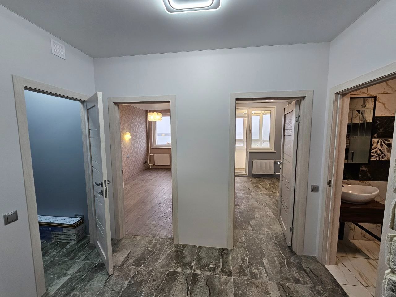 Wnętrze mieszkania po remoncie: widok na korytarz z otwartymi drzwiami do łazienki, pokoju i na balkon, szara marmurkowa podłoga, białe ściany i ościeżnice.