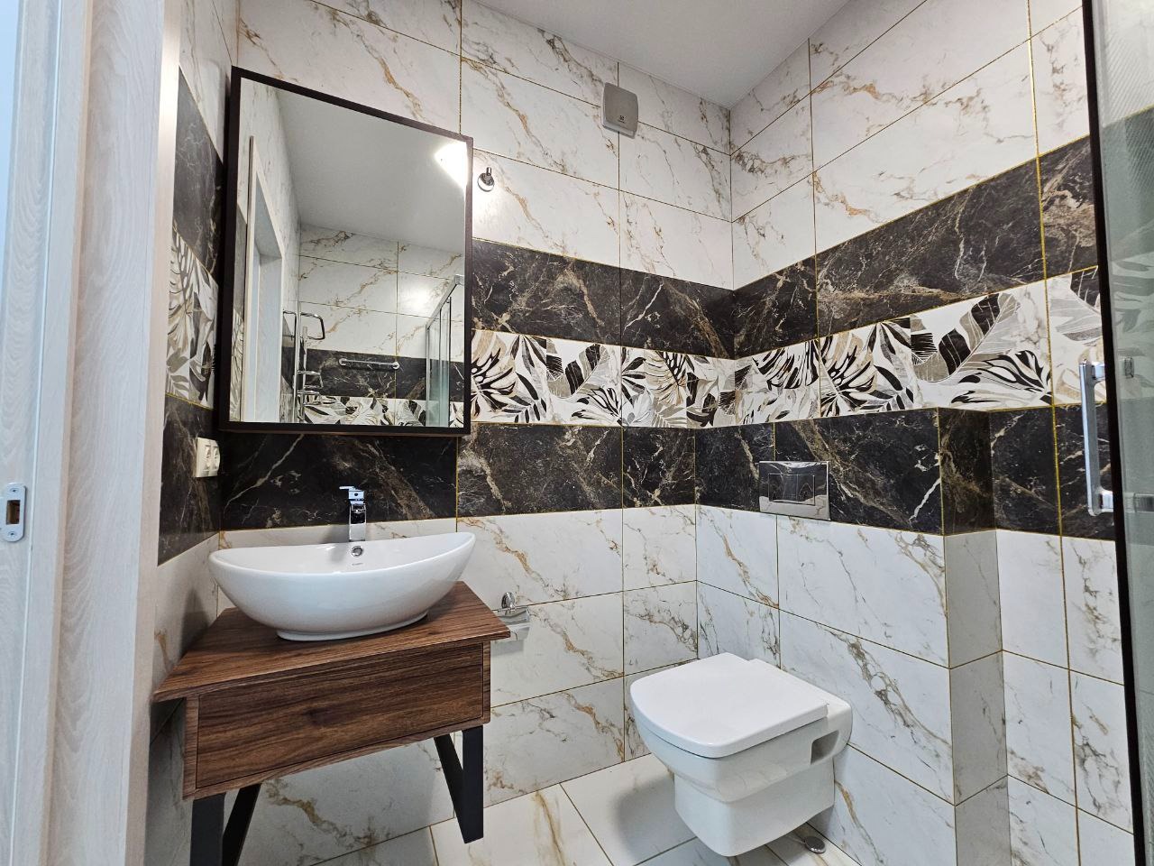 Nowoczesna łazienka z marmurowymi płytkami, umywalką nablatową na drewnianej szafce i dużym lustrem w czarnej ramie.