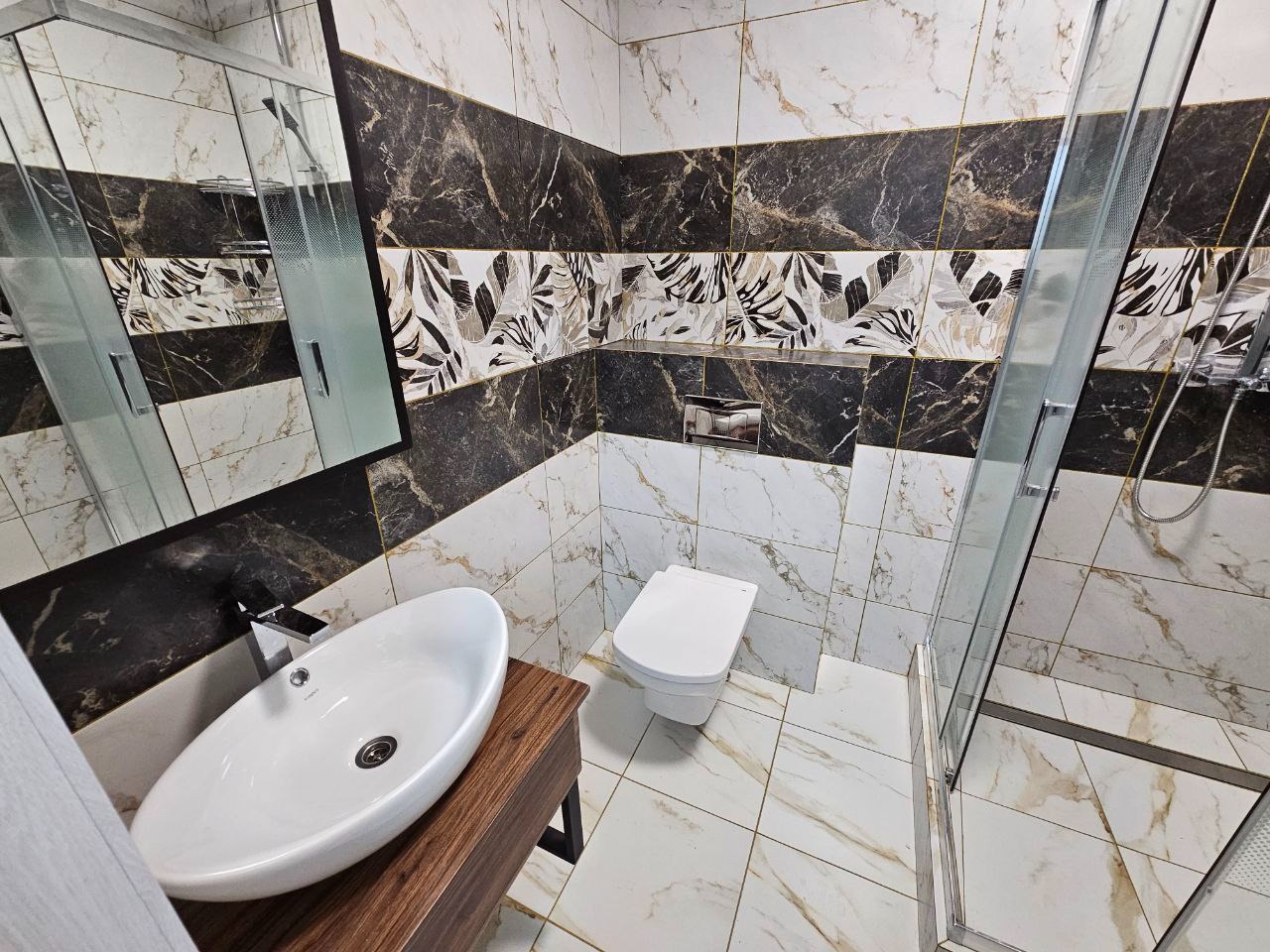 Nowoczesna łazienka z marmurowymi płytkami w odcieniach bieli i czerni, wzbogacona o dekoracyjny pas z motywem liści. Widoczna umywalka nablatowa na drewnianej szafce, toaleta oraz kabina...