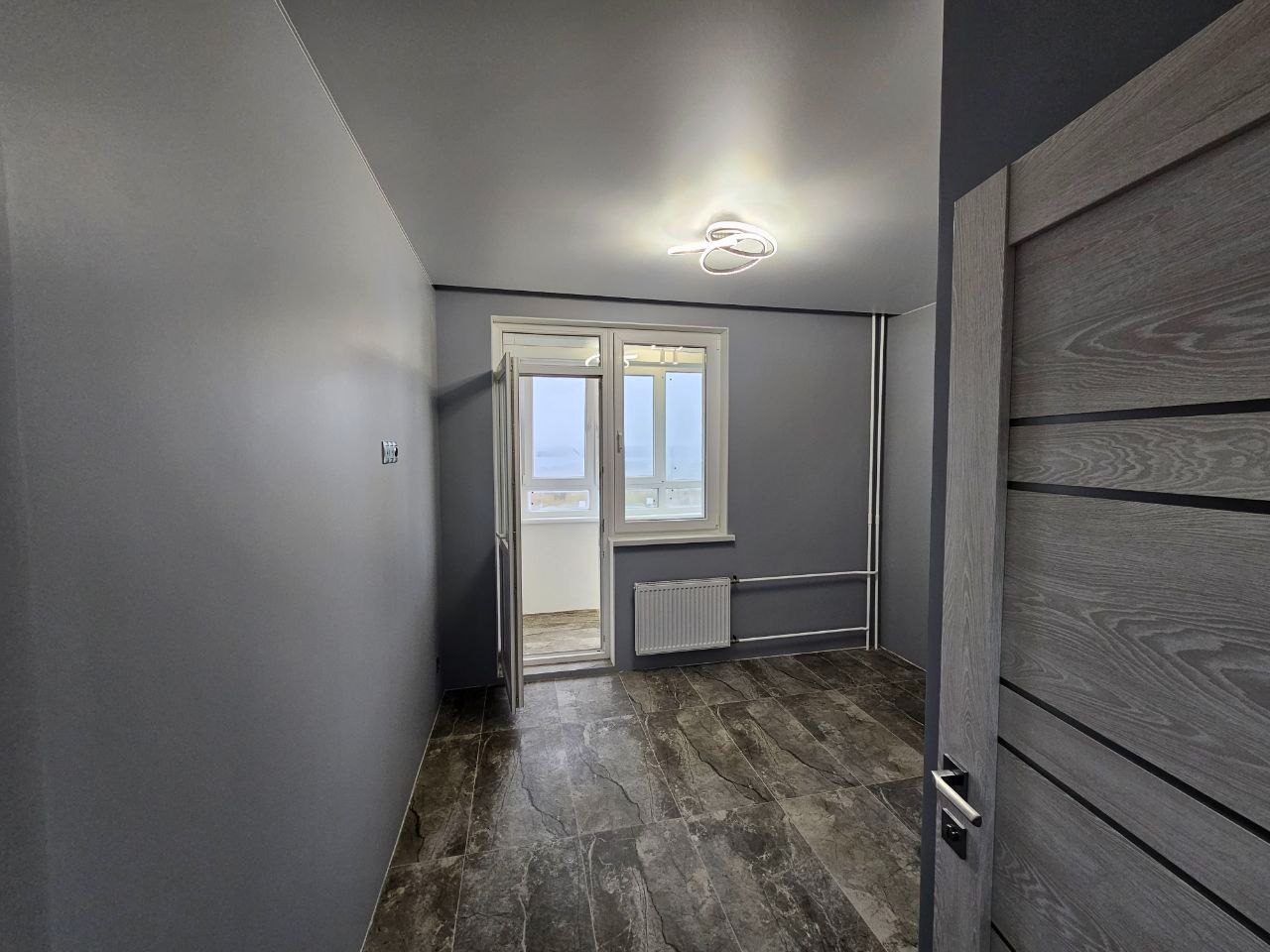 Szare wnętrze pokoju po remoncie z drzwiami z efektem drewna, marmurową podłogą, białym grzejnikiem i nowoczesną lampą sufitową.