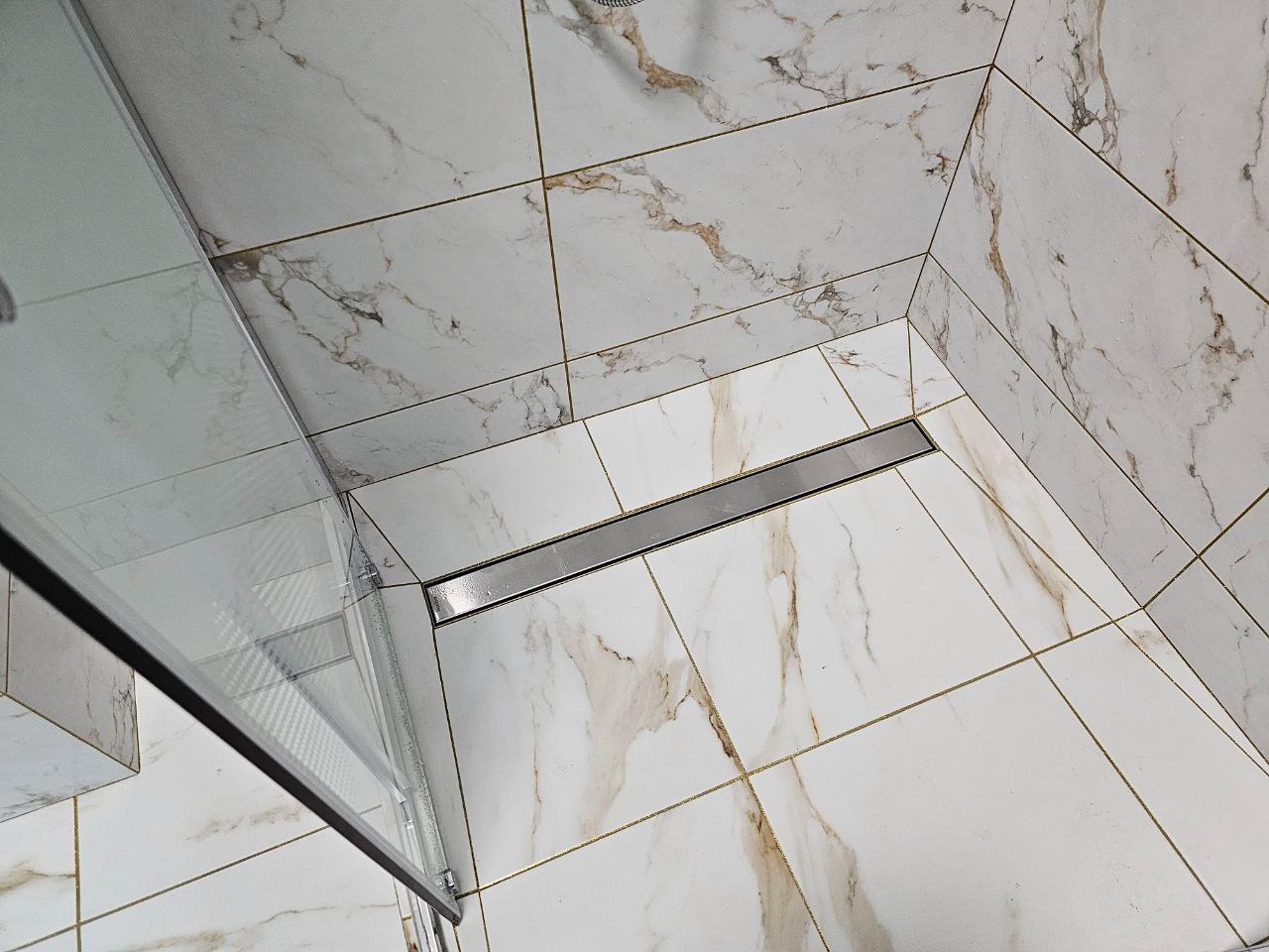 Nowoczesna kabina prysznicowa walk-in z odpływem liniowym, wykończona białymi płytkami imitującymi marmur z delikatnymi złotymi żyłkami. Widoczny fragment szklanej ścianki kabiny.