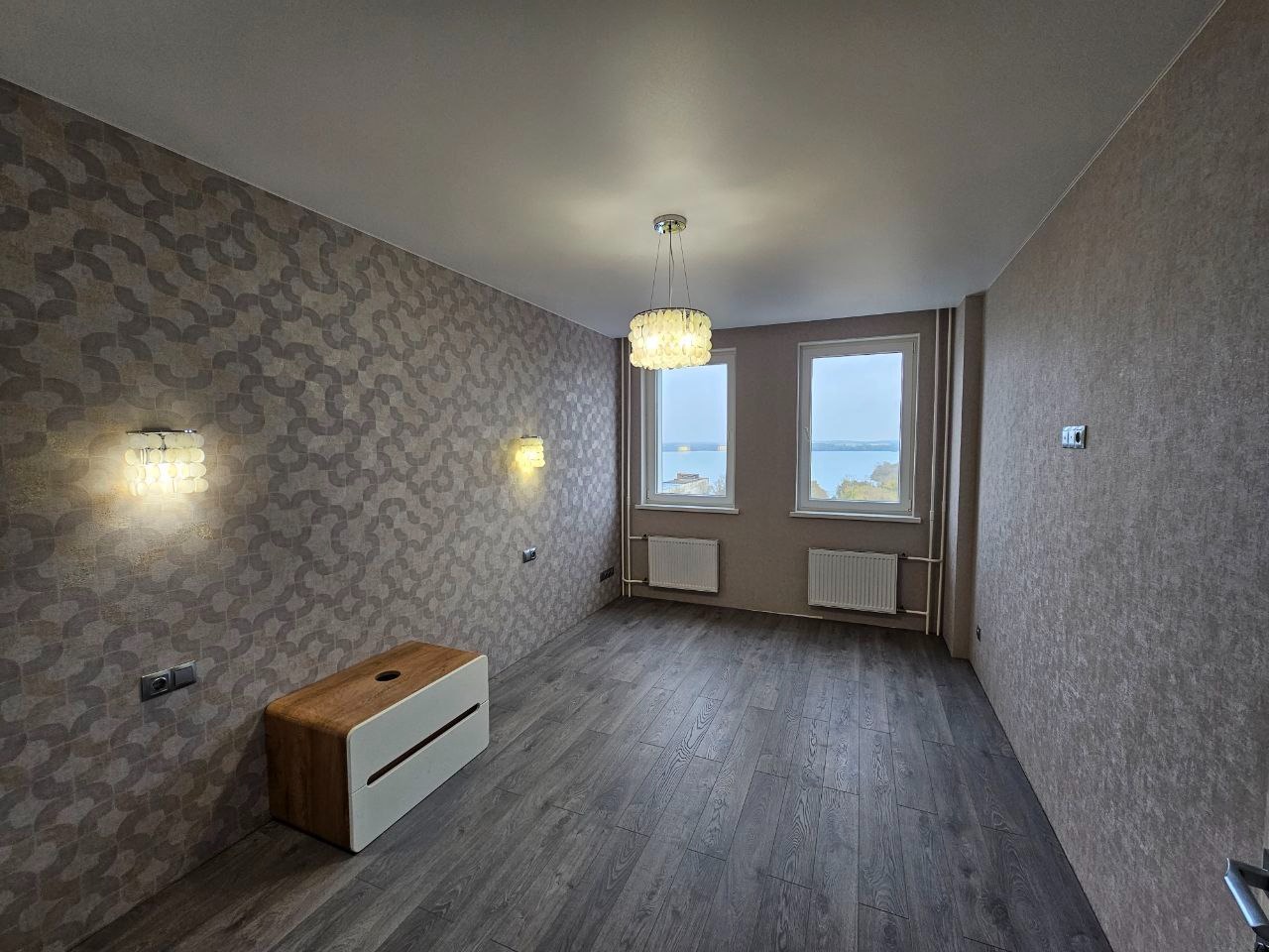 Wnętrze pokoju po remoncie: ciemne panele podłogowe, wzorzysta tapeta w odcieniach szarości i beżu, dekoracyjne lampy ścienne i sufitowa, widok na jezioro z okien.