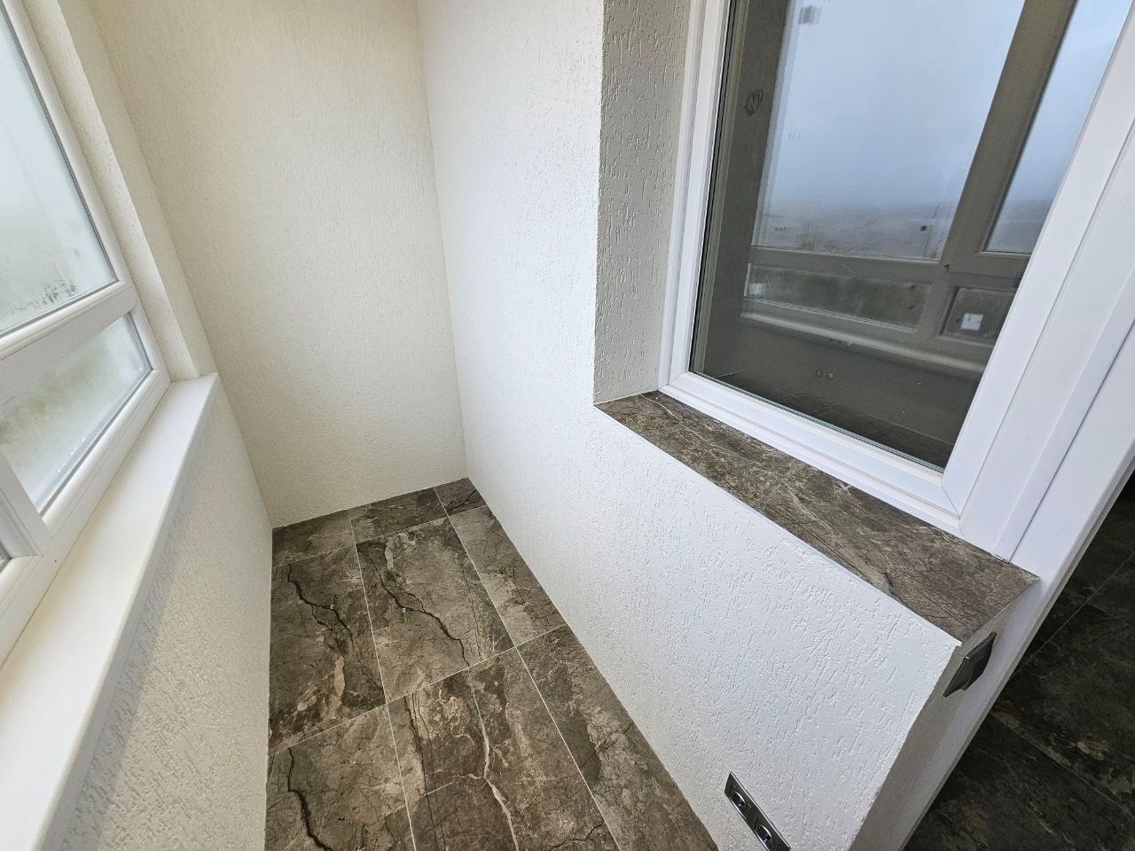 Wnętrze balkonu z szarobrązową marmurkową podłogą, białe ściany z fakturą baranka i białe okno z marmurowym parapetem.
