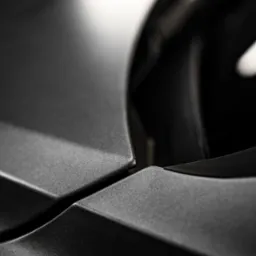 Detaliczne ujęcie matowej, czarnej karoserii samochodu w pobliżu reflektora, z widoczną fakturą lakieru i subtelną grą światła i cienia.