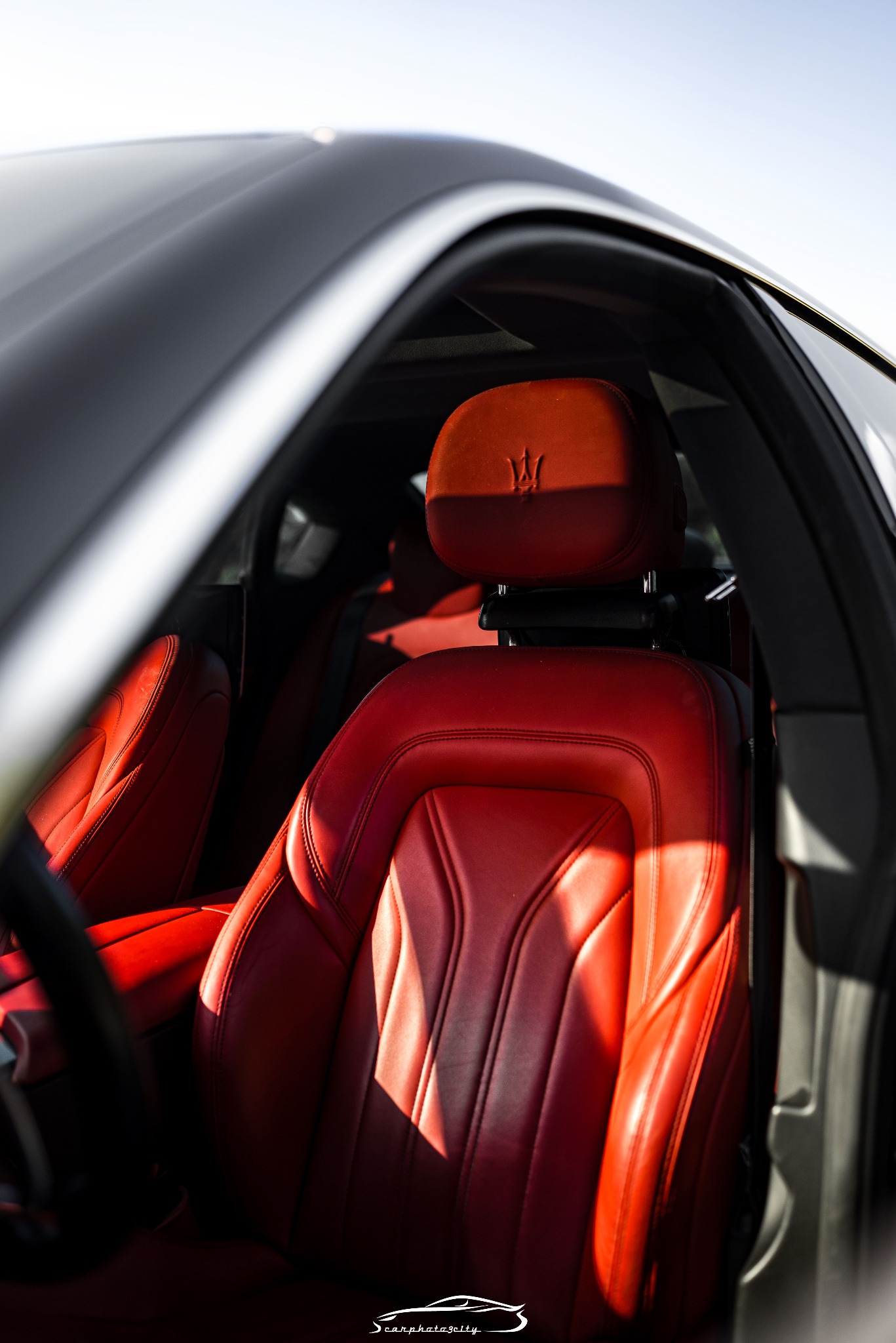 Czerwona skórzana tapicerka Maserati z wyraźnym logo na zagłówku, oświetlona promieniami słońca wpadającymi przez otwarte drzwi.