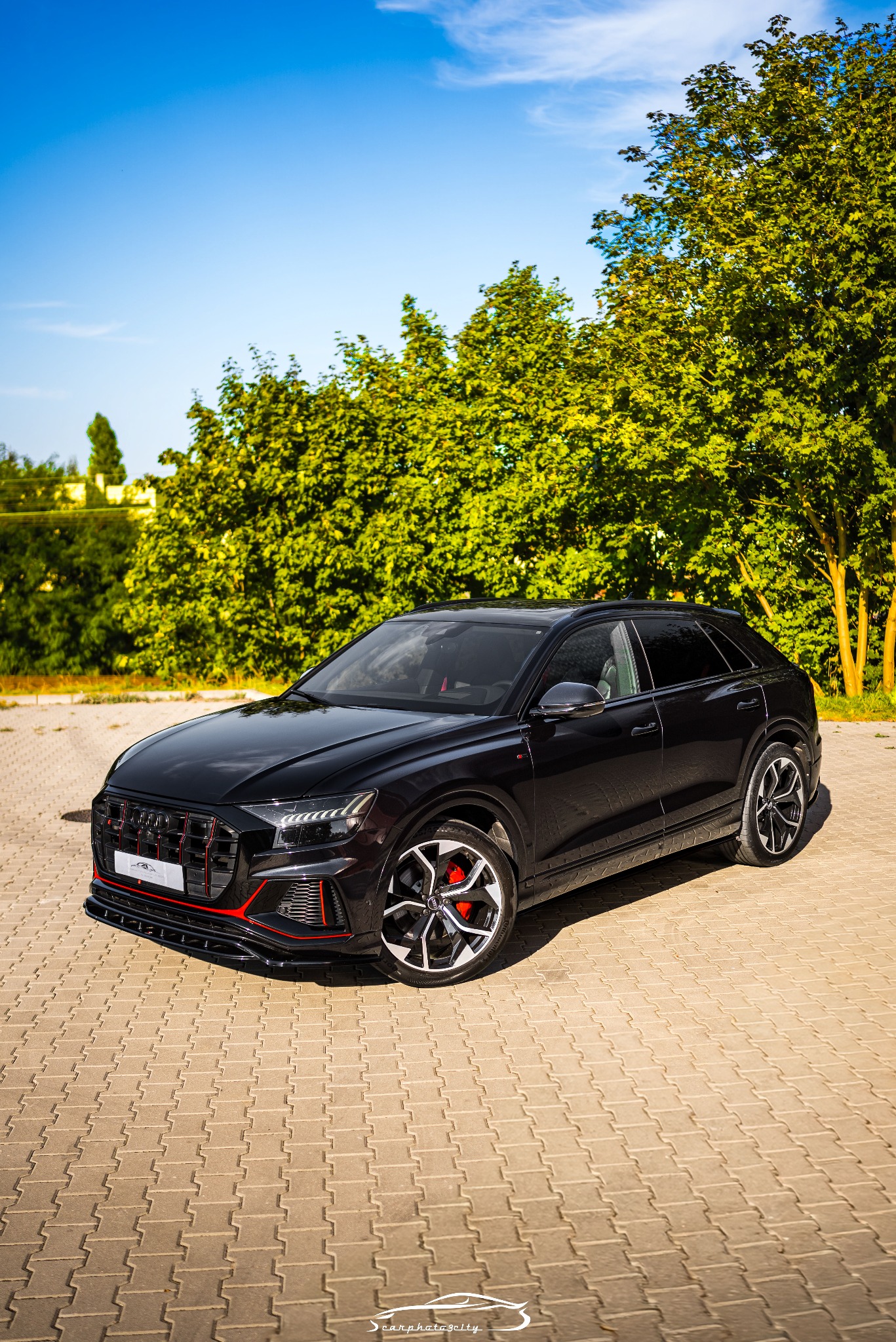 Czarny Audi Q8 po tuningu z czerwonymi akcentami, zaparkowany na kostce brukowej w słoneczny dzień, z widoczną zielenią w tle.