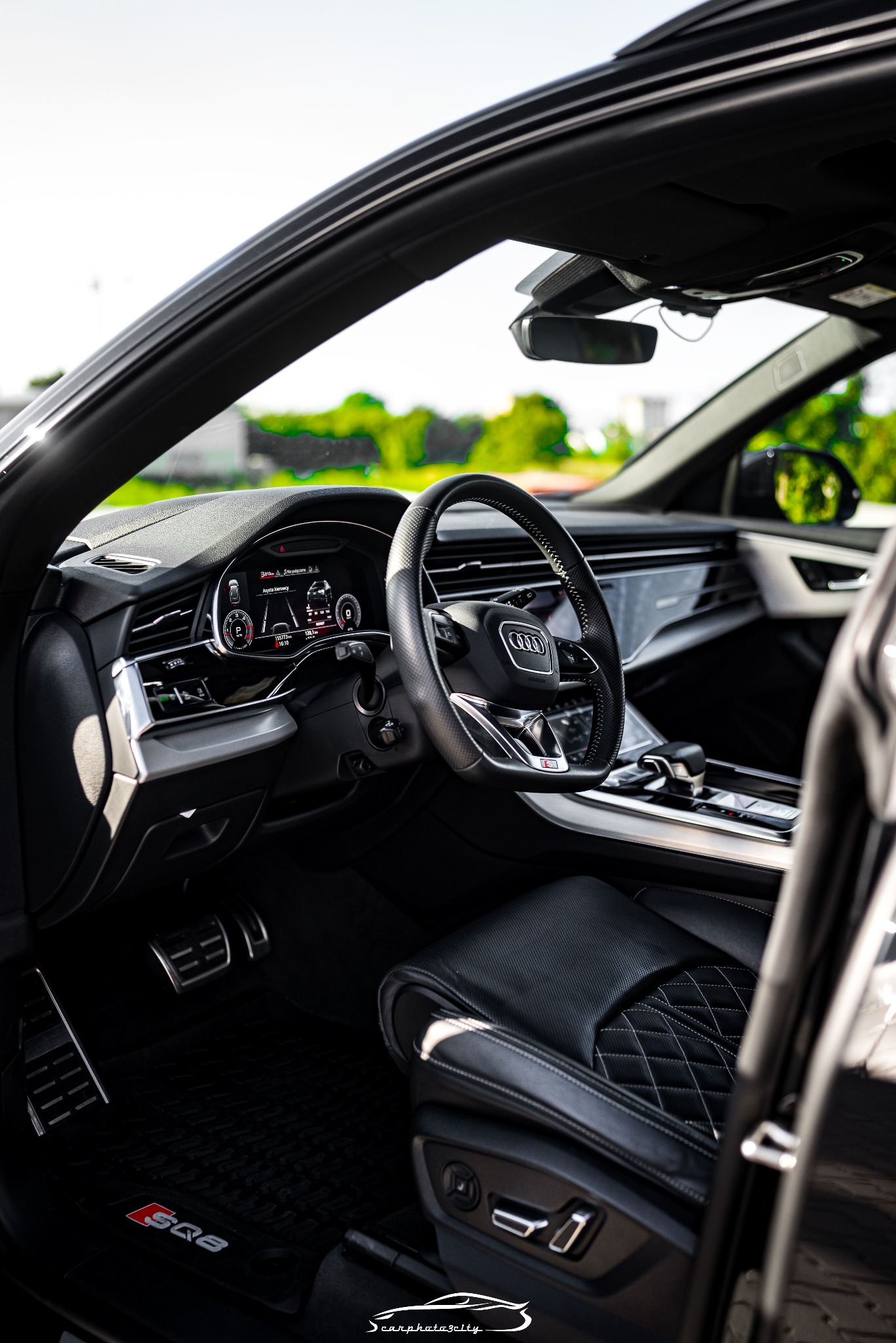Wnętrze luksusowego Audi Q8 z czarną skórzaną tapicerką, widok z zewnątrz przez otwarte drzwi, deska rozdzielcza z cyfrowym wyświetlaczem i logo S Q8 na dywaniku.