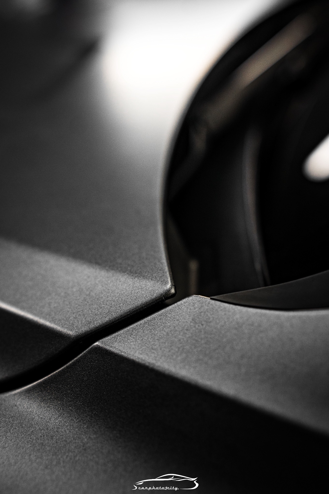 Detaliczne ujęcie matowej, czarnej karoserii samochodu w pobliżu reflektora, z widoczną fakturą lakieru i subtelną grą światła i cienia.