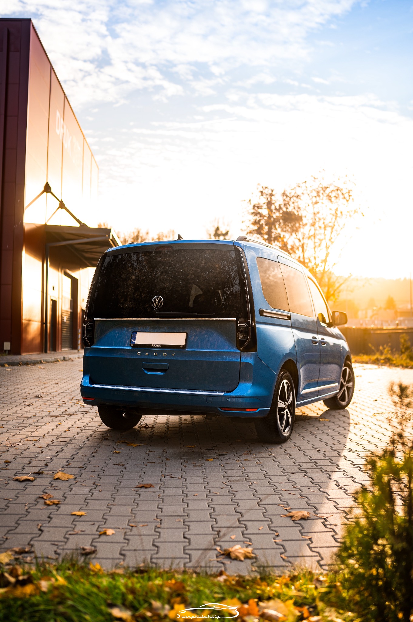 Błękitny Volkswagen Caddy zaparkowany na brukowanym placu, jesienne liście, zachodzące słońce odbija się w szybach