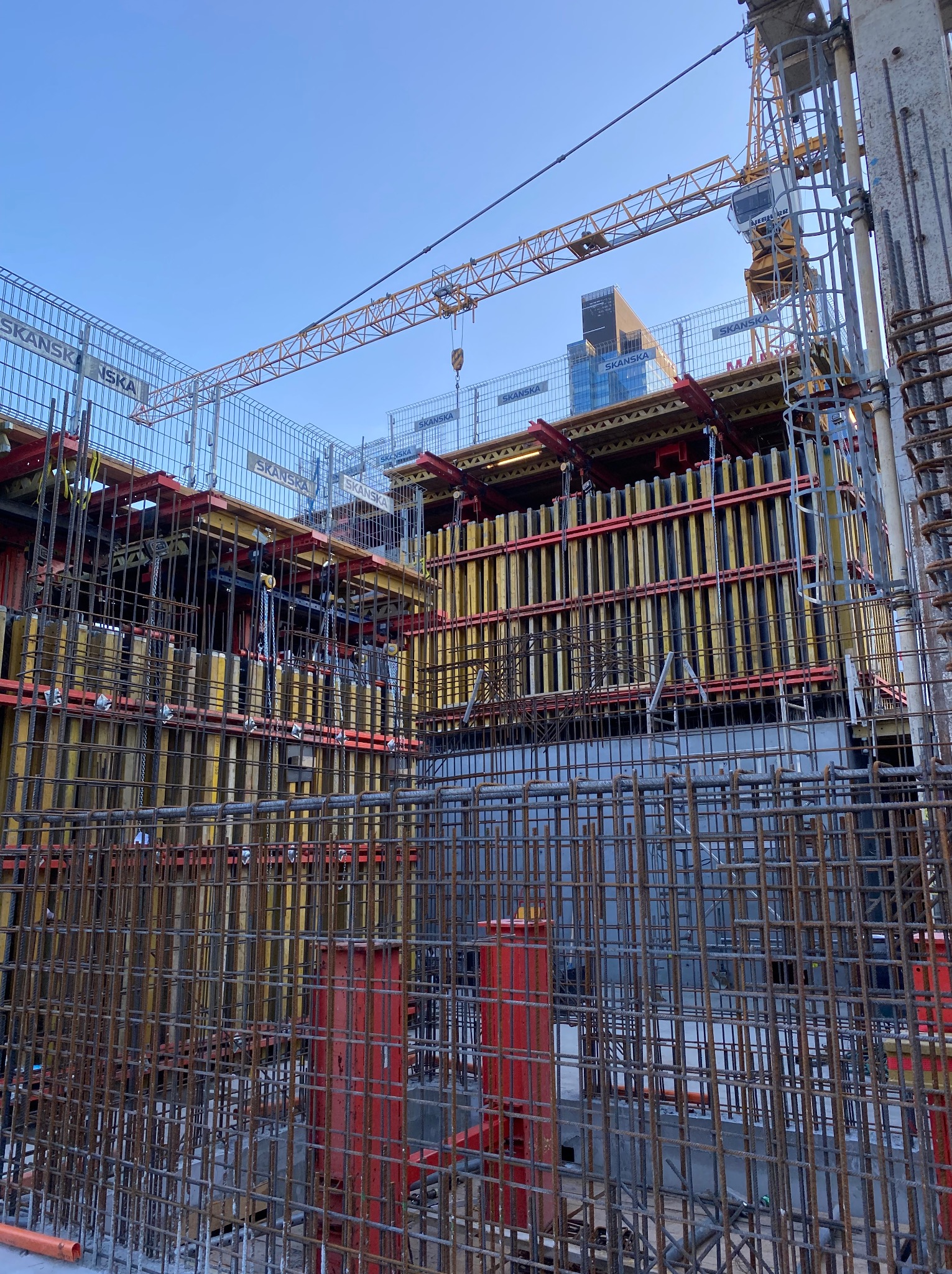 Konstrukcja zbrojeniowa z widocznymi elementami szalunków i żurawiem wieżowym w tle, wczesny etap budowy budynku z elementami logo Skanska na szalunkach.