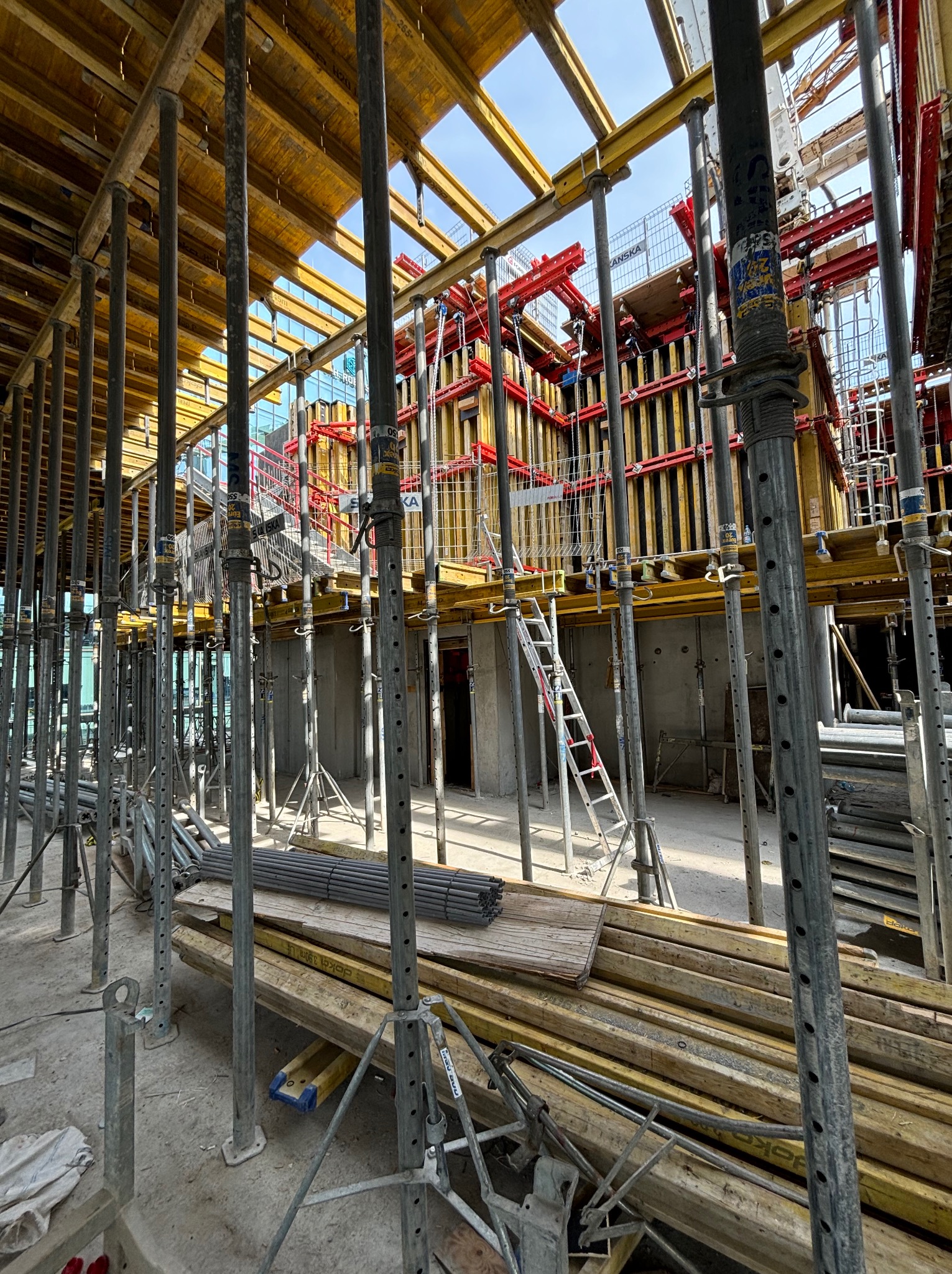Wnętrze placu budowy z widocznymi stalowymi podporami, szalunkami stropowymi i pionowymi, drabina aluminiowa oparta o ścianę, w tle konstrukcja budynku w trakcie wznoszenia.