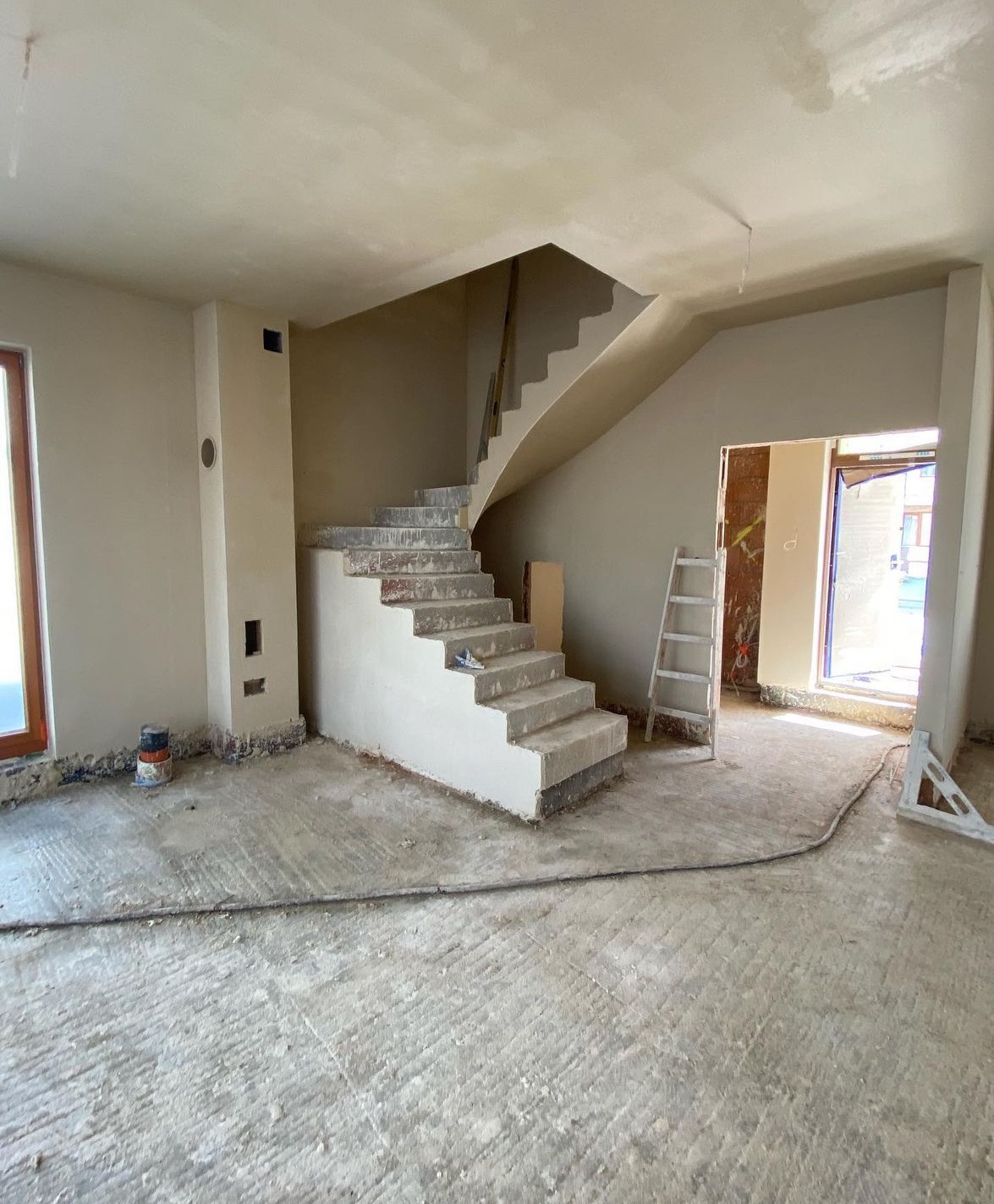 Wnętrze domu w trakcie remontu: surowe schody betonowe, świeżo otynkowane ściany, widoczna drabina oparta o ścianę przy wejściu, ślady farby na stopniach schodów i niedokończona podłoga.