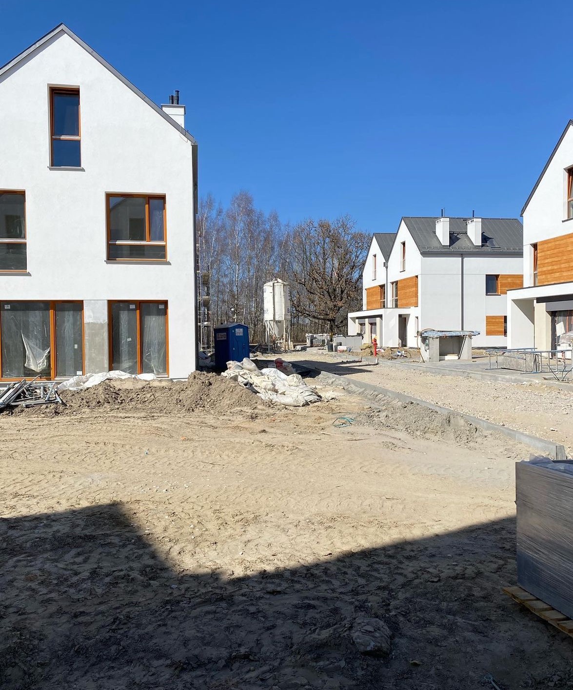 Osiedle nowych domów jednorodzinnych w trakcie budowy, widoczne białe elewacje z drewnianymi akcentami, teren przygotowany pod trawnik, worki z materiałami budowlanymi i przenośna toaleta w tle.