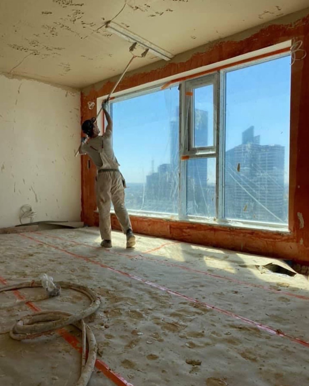 Pracownik w kombinezonie wyrównuje sufit szpachlą w pokoju z dużym oknem i widokiem na miasto, podłoga zabezpieczona folią, widoczne ślady prac remontowych.