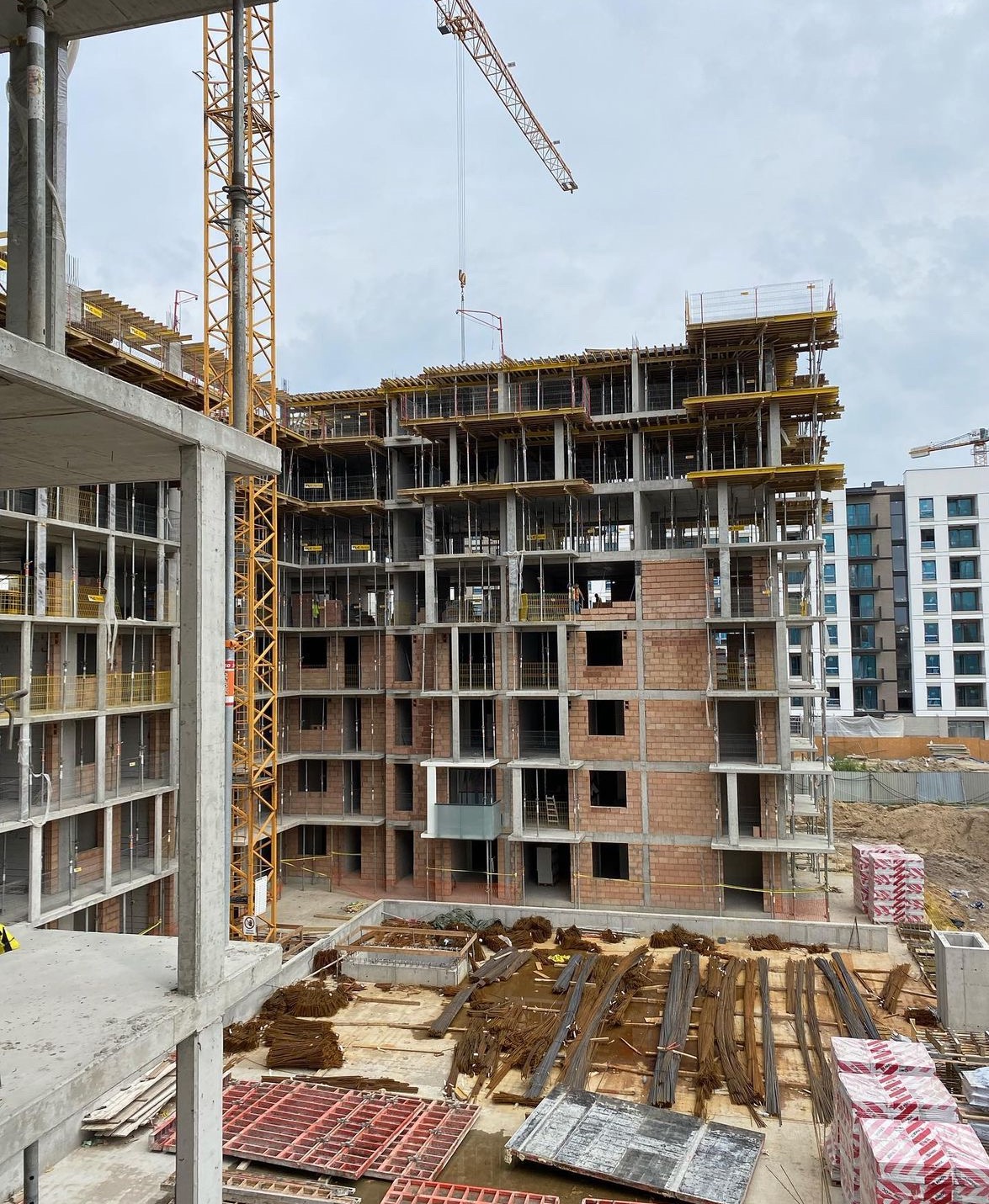 Wysoki budynek w stanie surowym otwartym, z widocznymi elementami konstrukcyjnymi, rusztowaniami i żurawiami na placu budowy, sfotografowany pod pochmurnym niebem.