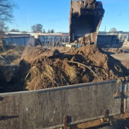 Jakub Wawrzynowicz - Ładowarka nabiera ziemię z wykopu i wrzuca ją do skrzyni ciężarówki. W tle budynki gospodarcze i błękitne niebo.