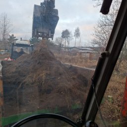 Jakub Wawrzynowicz - Koparka nabiera ziemię z wykopu, widok z kabiny operatora. W tle zabudowania i drzewa.