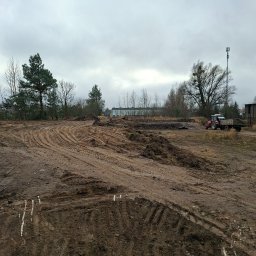 Jakub Wawrzynowicz - Przygotowany teren pod budowę z widocznymi śladami opon i traktorem w tle, pochmurne niebo.