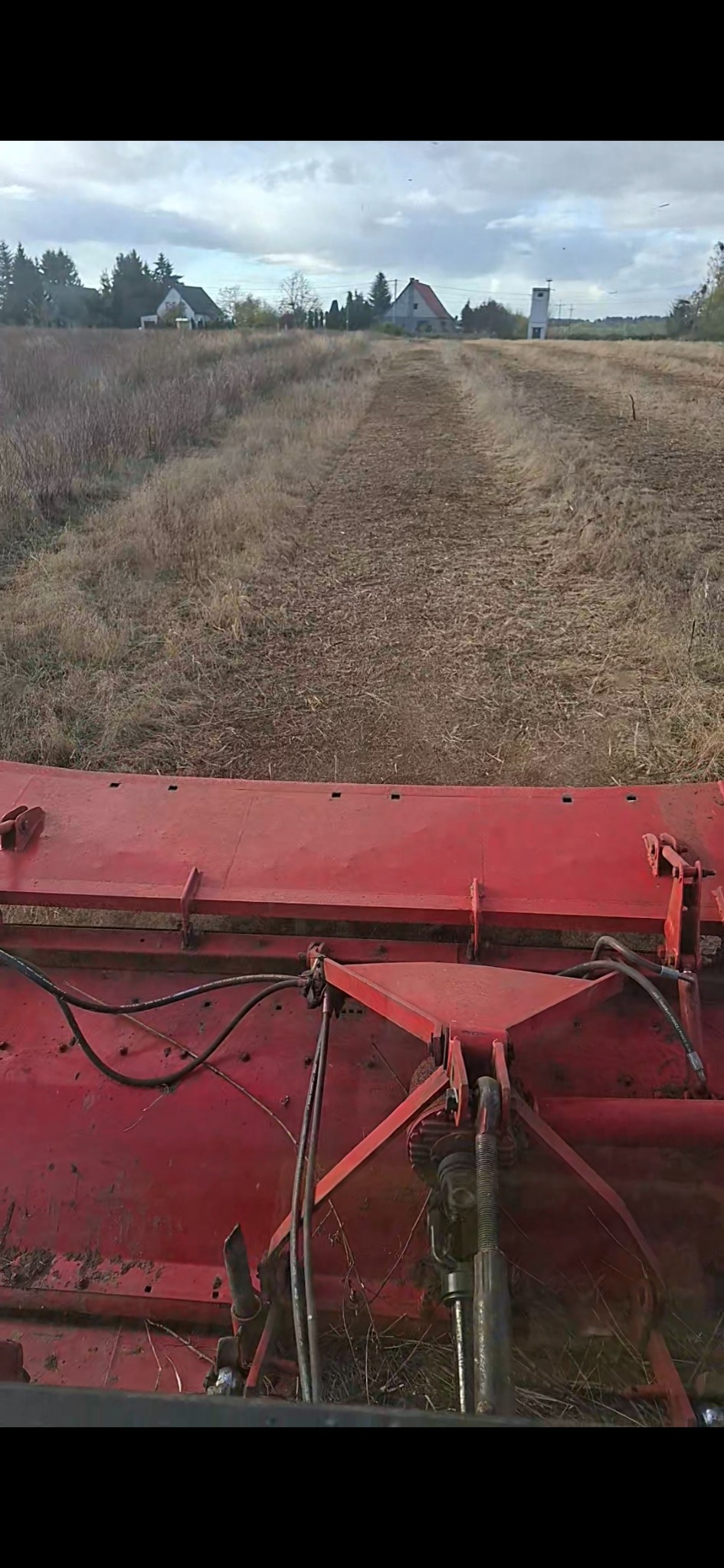 Czerwona maszyna rolnicza w trakcie pracy na polu, widok z perspektywy operatora, w tle zabudowania wiejskie i pochmurne niebo. Ślady opon na ziemi.