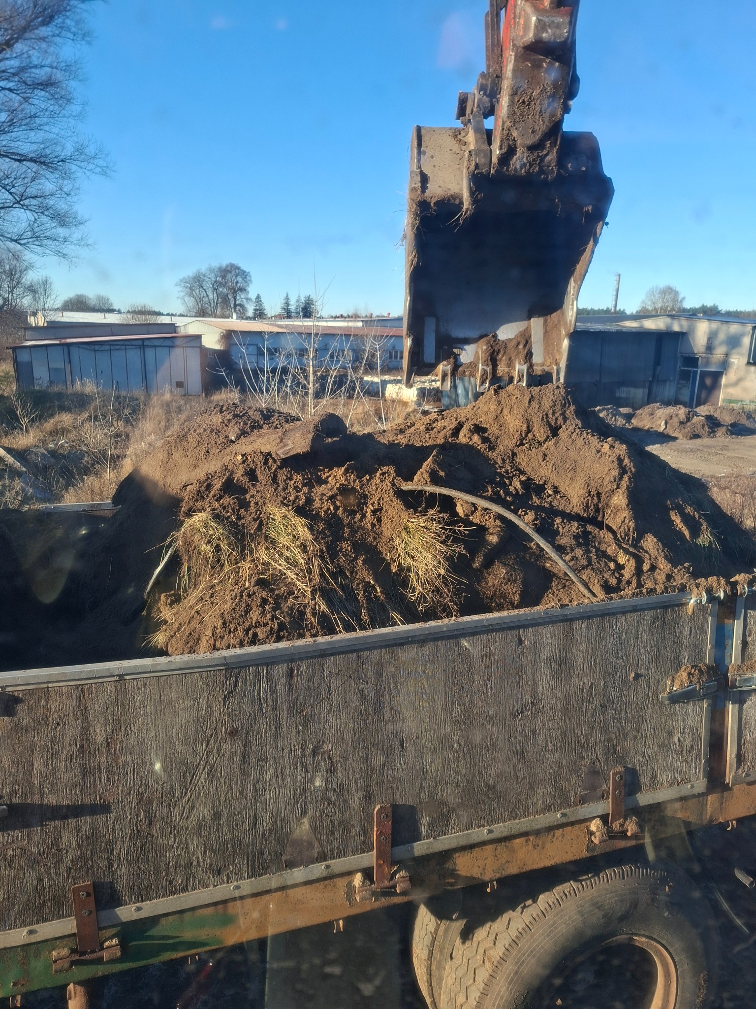 Ładowarka nabiera ziemię z wykopu i wrzuca ją do skrzyni ciężarówki. W tle budynki gospodarcze i błękitne niebo.