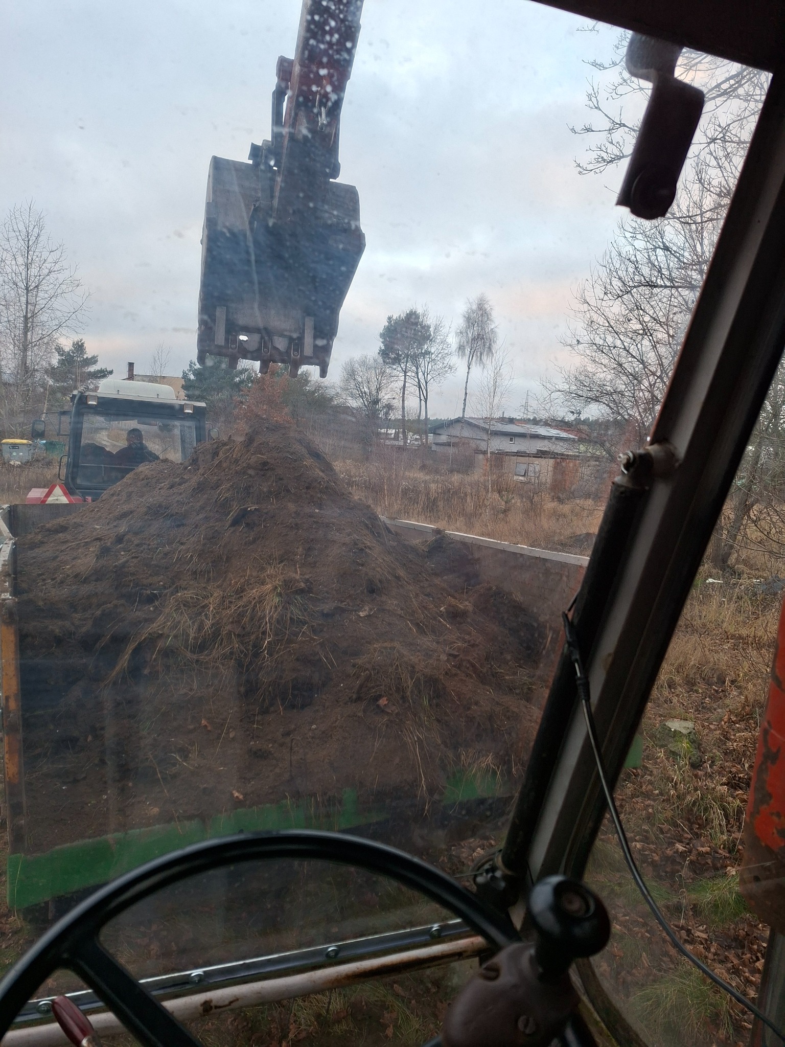 Koparka nabiera ziemię z wykopu, widok z kabiny operatora. W tle zabudowania i drzewa.