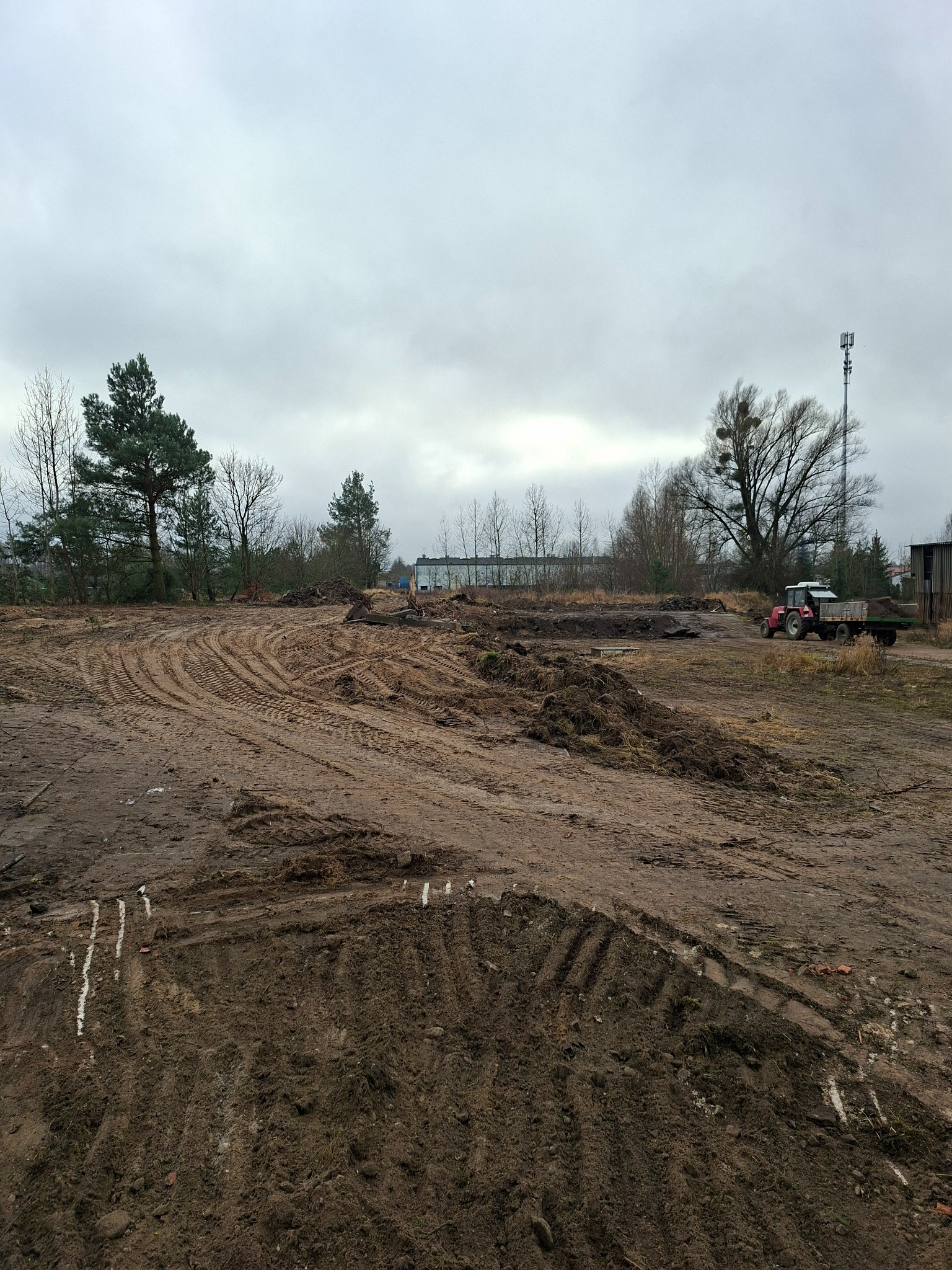 Przygotowany teren pod budowę z widocznymi śladami opon i traktorem w tle, pochmurne niebo.