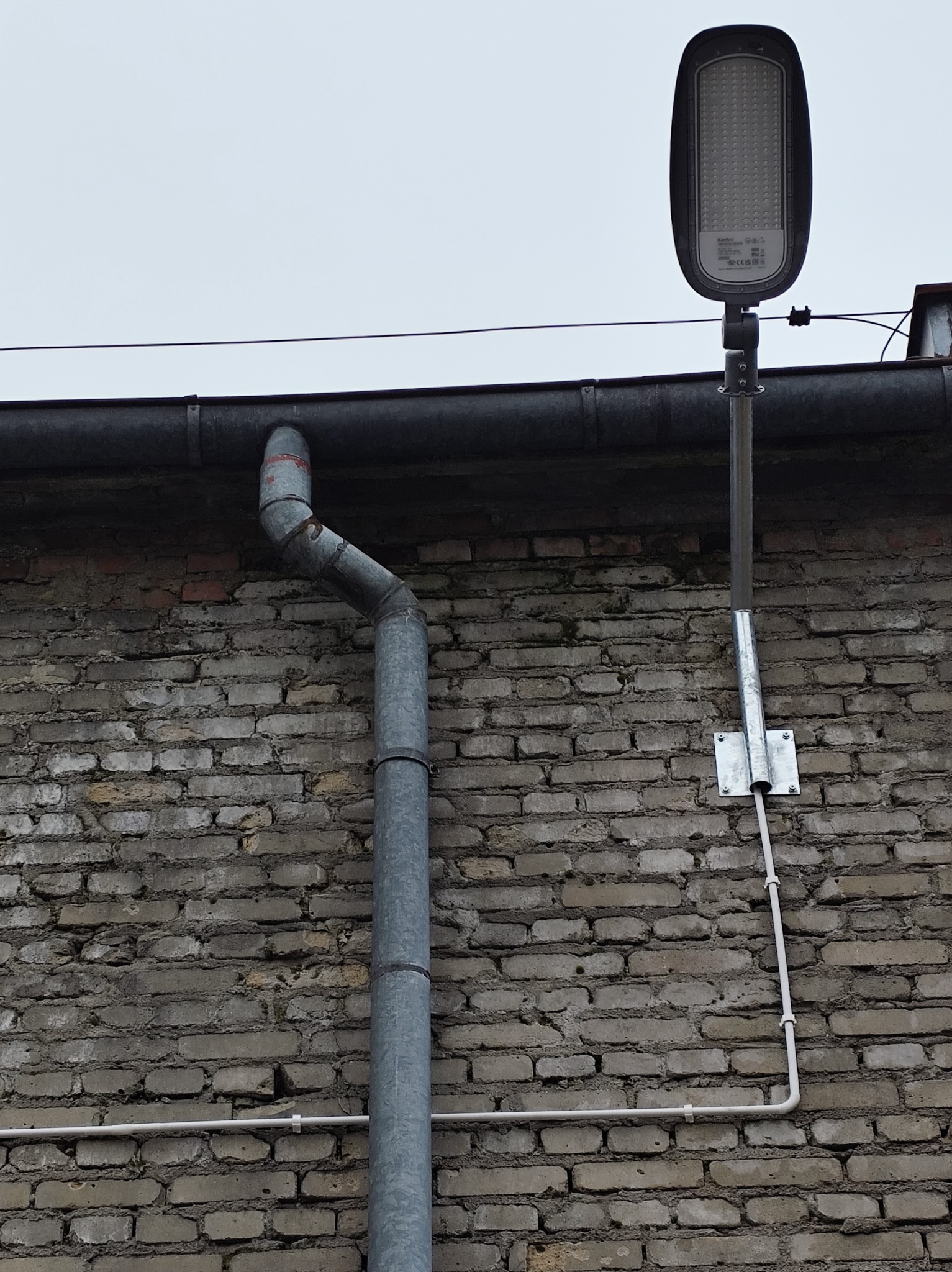 Zamontowana lampa LED na szarym słupie przy ceglanej ścianie budynku z rynną. Widoczne przewody elektryczne. Ujęcie z dołu, pochmurne niebo w tle.