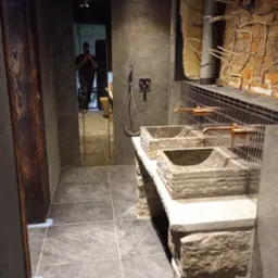 Nowoczesna łazienka z dwoma kamiennymi umywalkami i miedzianymi kranami. W tle lustro z odbiciem osoby robiącej zdjęcie. Widoczna instalacja na ścianie z cegły.