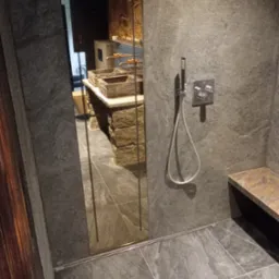 Nowoczesna łazienka z kamienną ścianą i kabiną prysznicową. Widoczny panel sterujący prysznicem i kamienna ławeczka. W lustrze odbija się umywalka z kamienia.