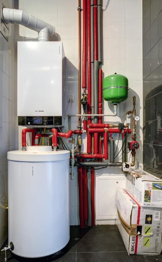 Biały piec grzewczy Viessmann z zasobnikiem ciepłej wody, podłączony do instalacji z rur w izolacji czerwonej i szarej, zielony zbiornik wyrównawczy, na tle białych płytek i ciemnej podłogi, w rogu...