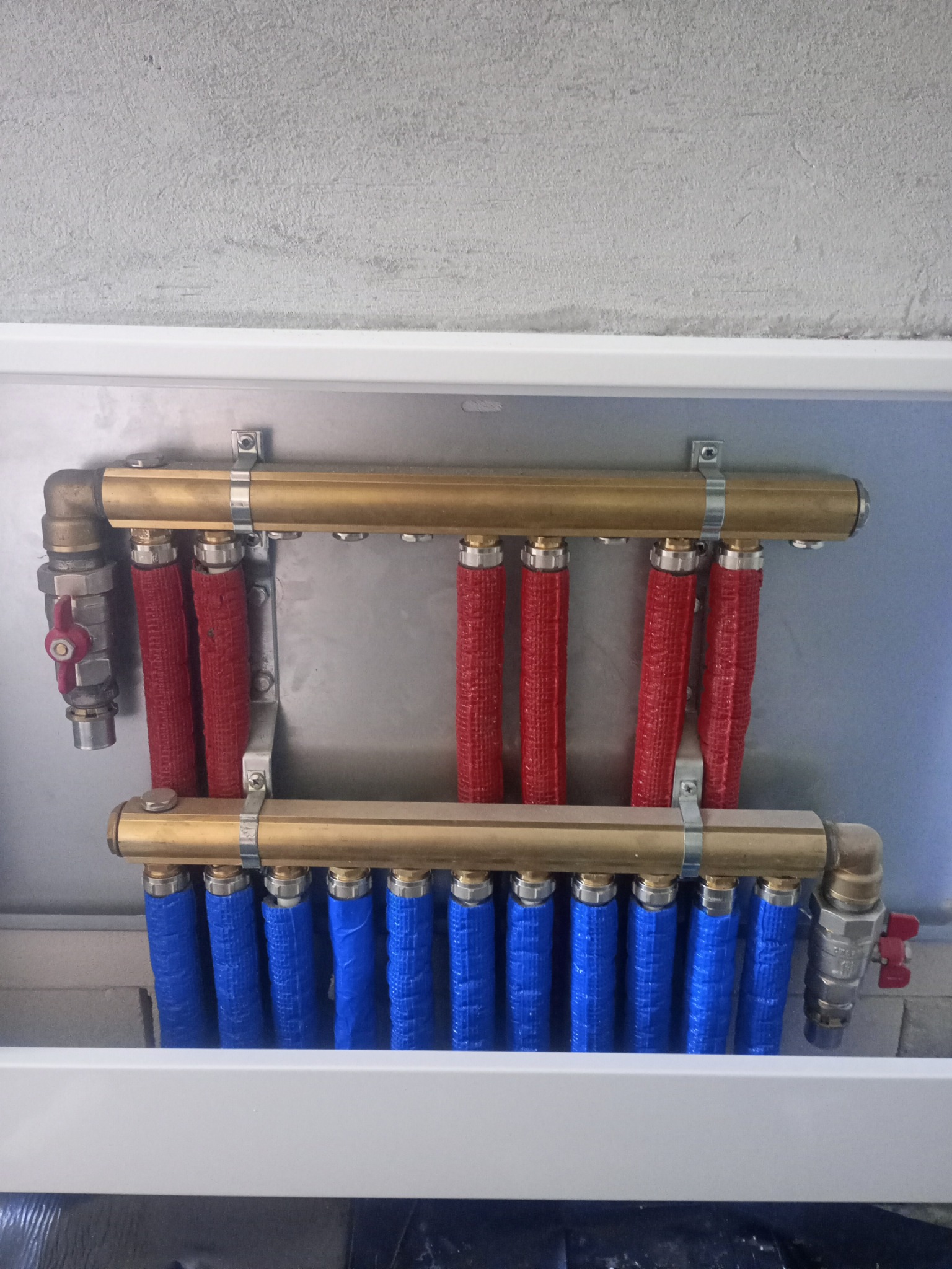 Rozdzielacz ogrzewania podłogowego z rurami w izolacji czerwonej i niebieskiej, zamontowany w białej szafce, widoczny fragment ściany z surowego betonu