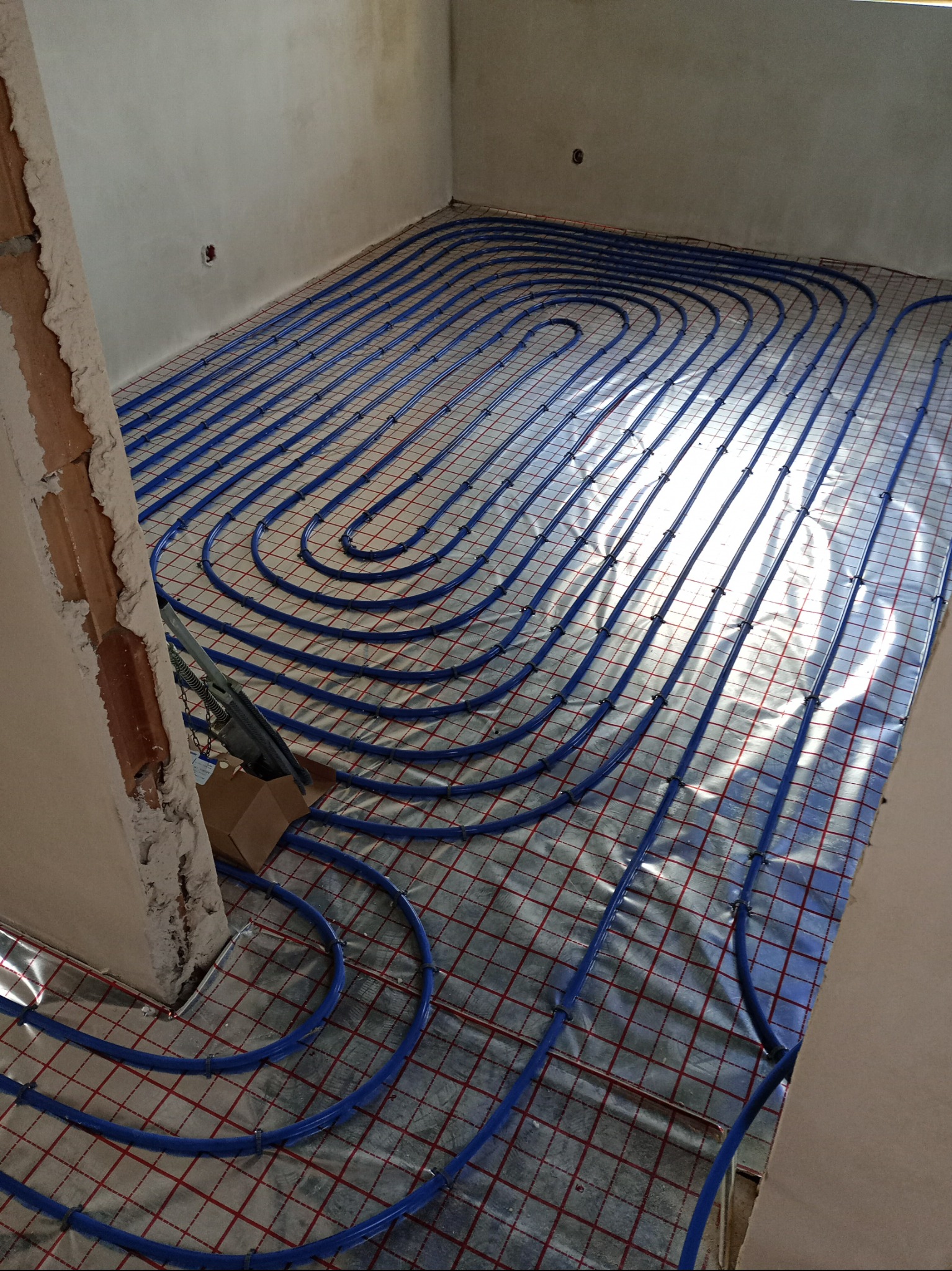 Niebieskie rury ogrzewania podłogowego ułożone w pętle na siatce z folią aluminiową w surowym pomieszczeniu, widoczny fragment ściany w stanie deweloperskim.