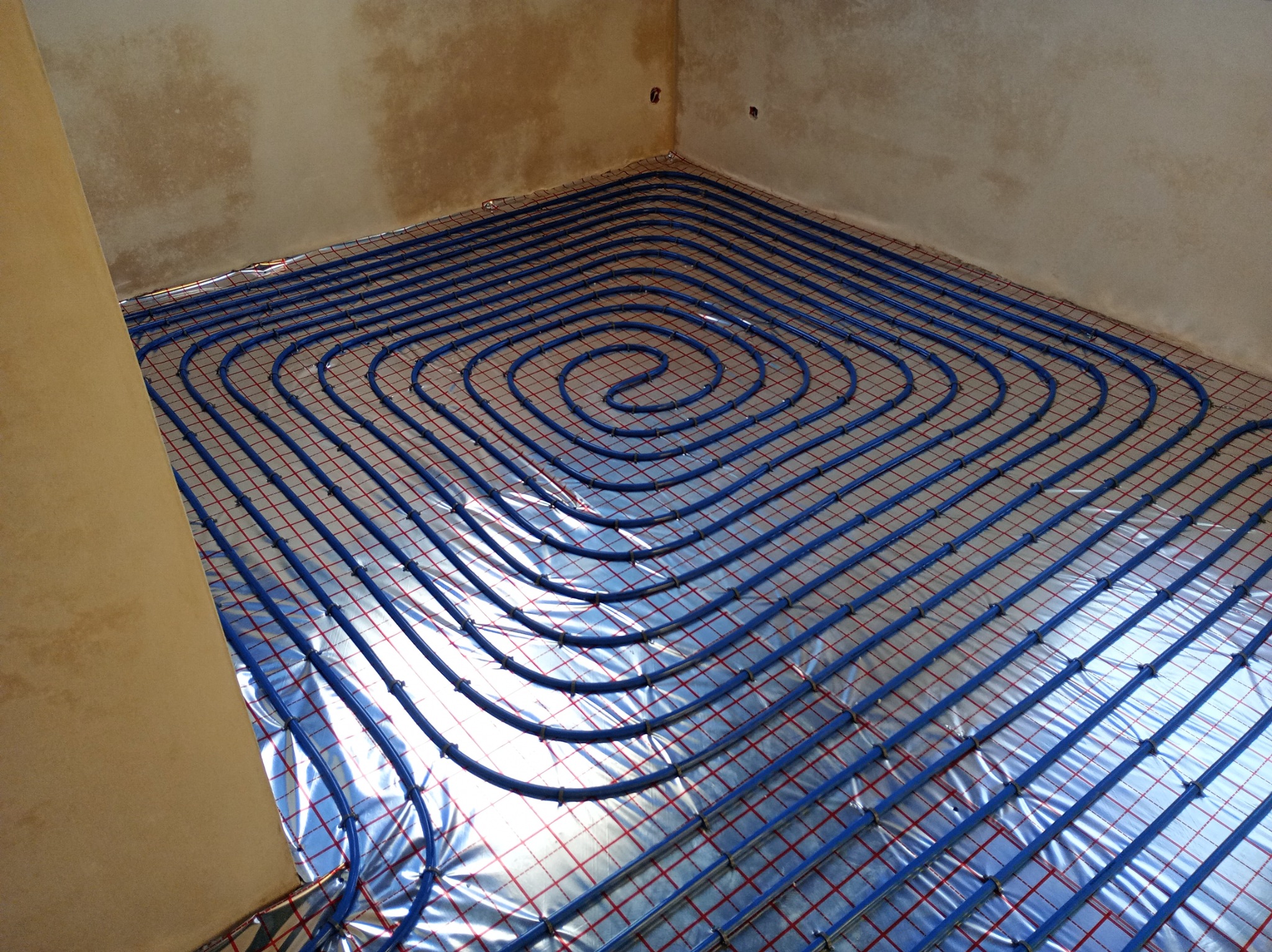 Niebieskie rury ogrzewania podłogowego ułożone spiralnie na srebrnej folii z czerwoną siatką na podłodze w surowym pomieszczeniu.
