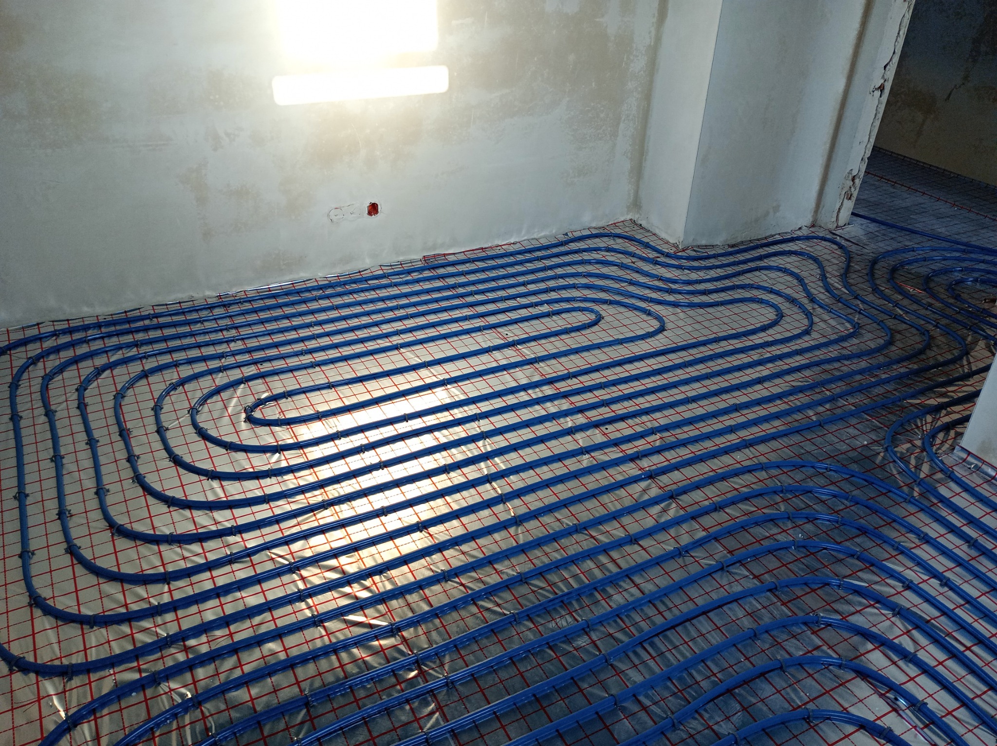 Niebieskie rury ogrzewania podłogowego ułożone na folii z czerwoną siatką w surowym wnętrzu, widoczne fragmenty ścian w stanie deweloperskim.